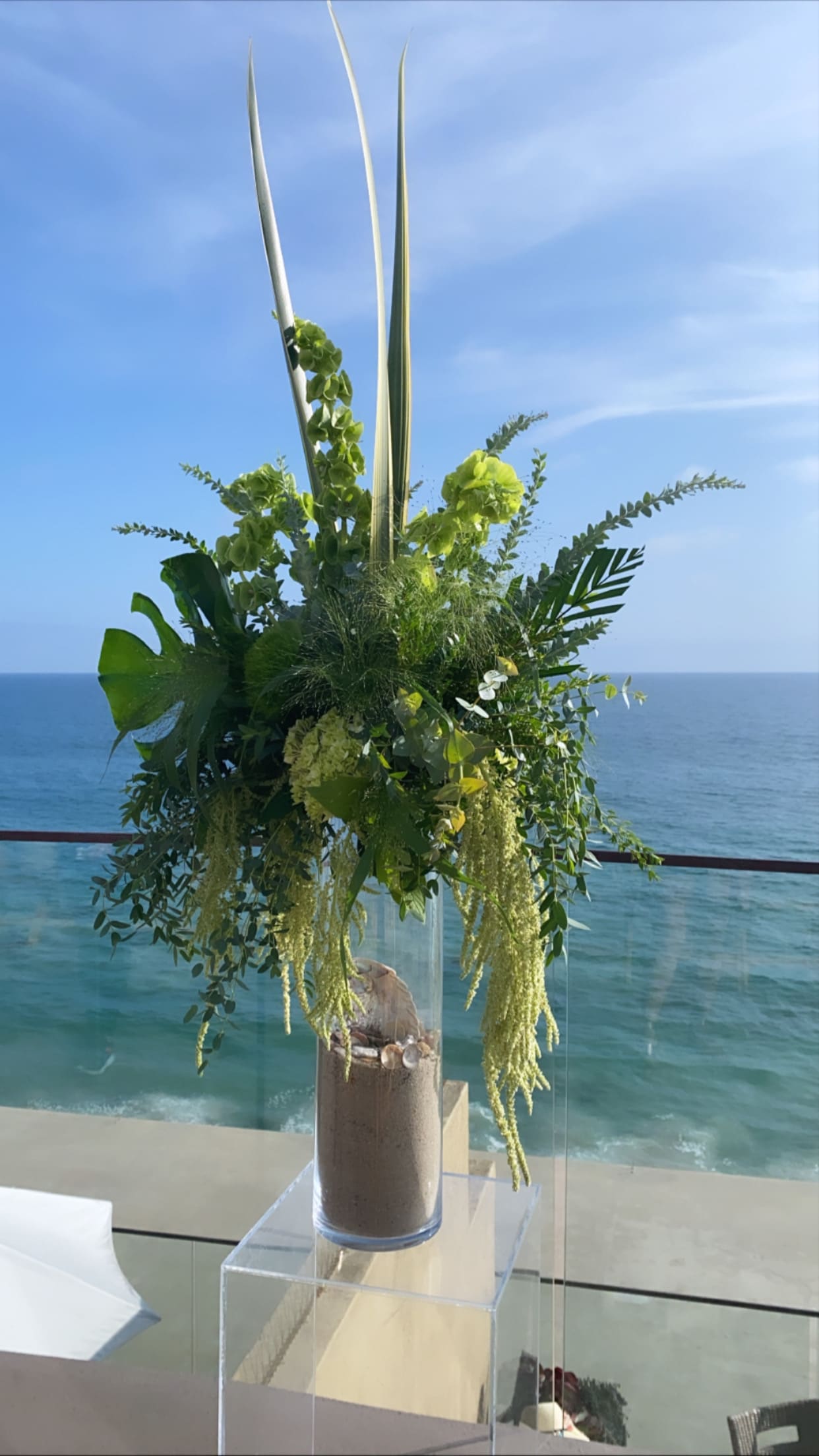 Tall green floral arrangement in a clear vase by the ocean
