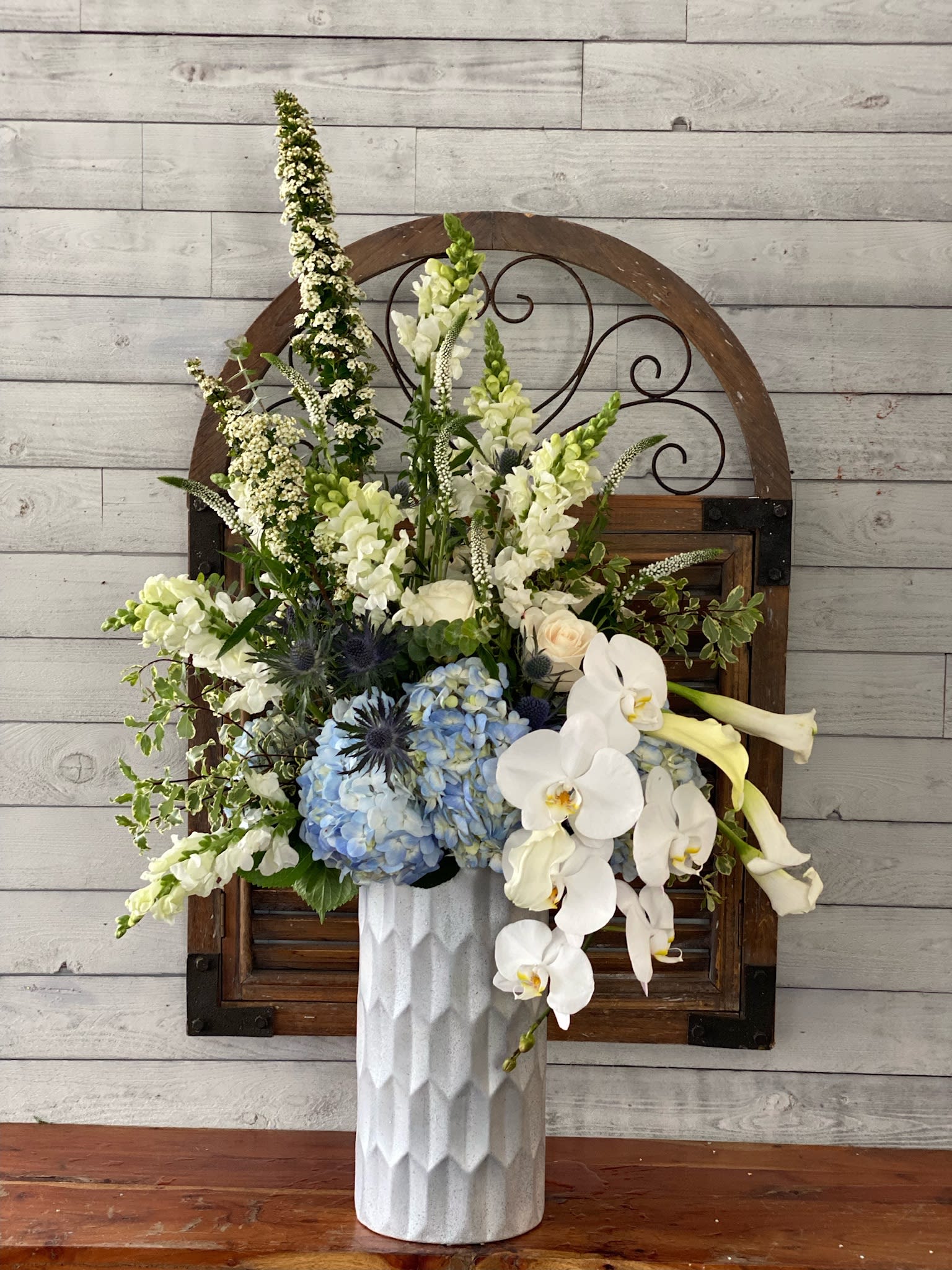 Tall floral arrangement with white orchids, blue hydrangeas, and pale blooms in a white vase