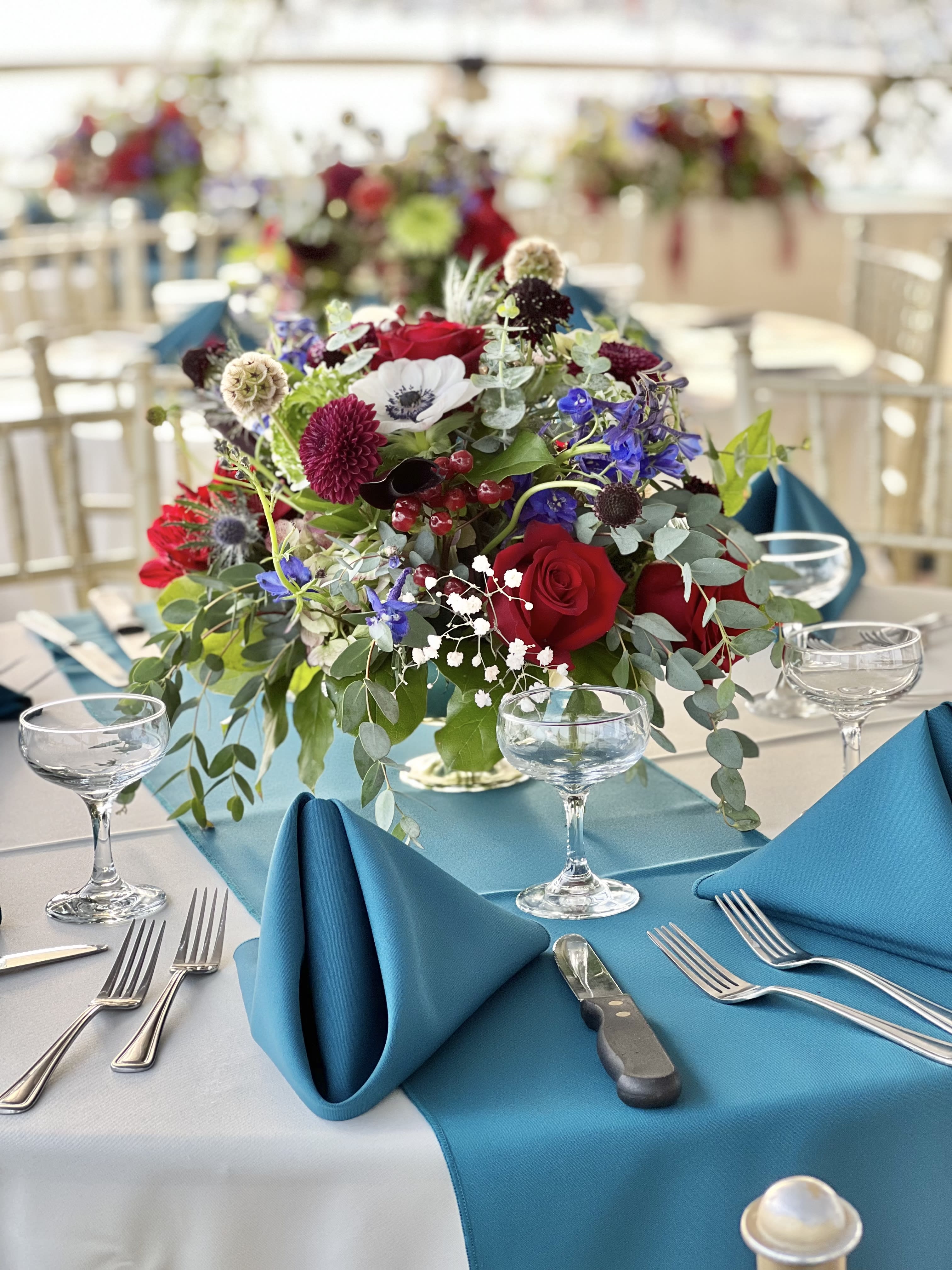 Low centerpiece with red roses, blue flowers, and greenery on a banquet table