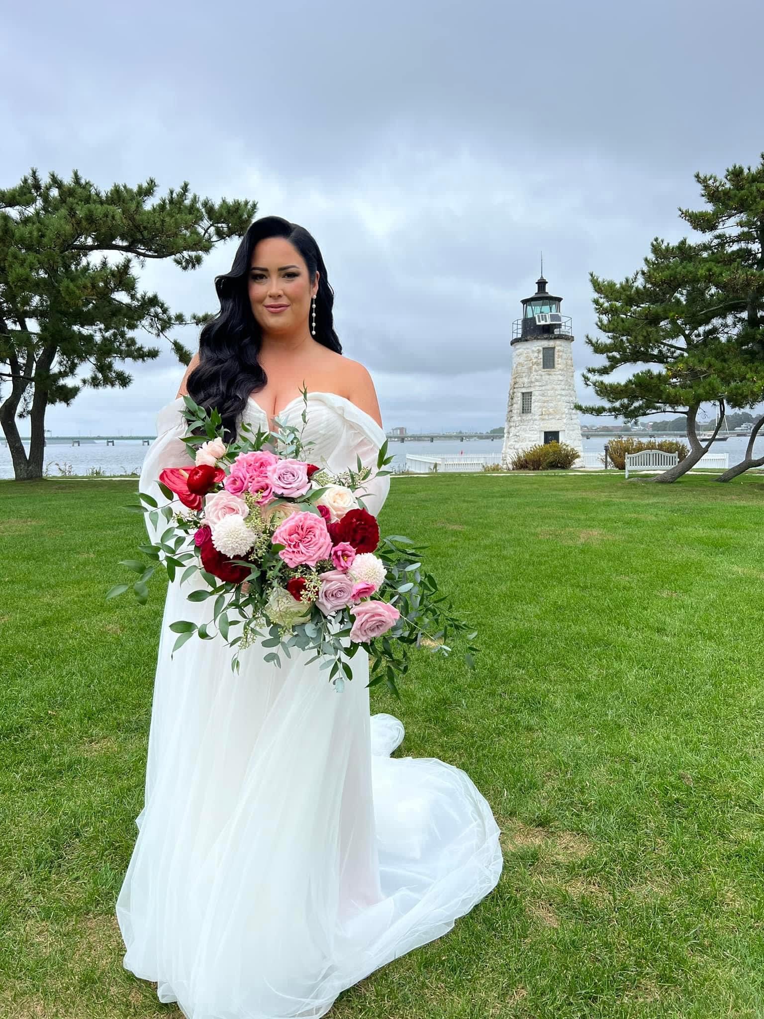 Bride holding a pink and red bouquet outdoors near a lighthouse