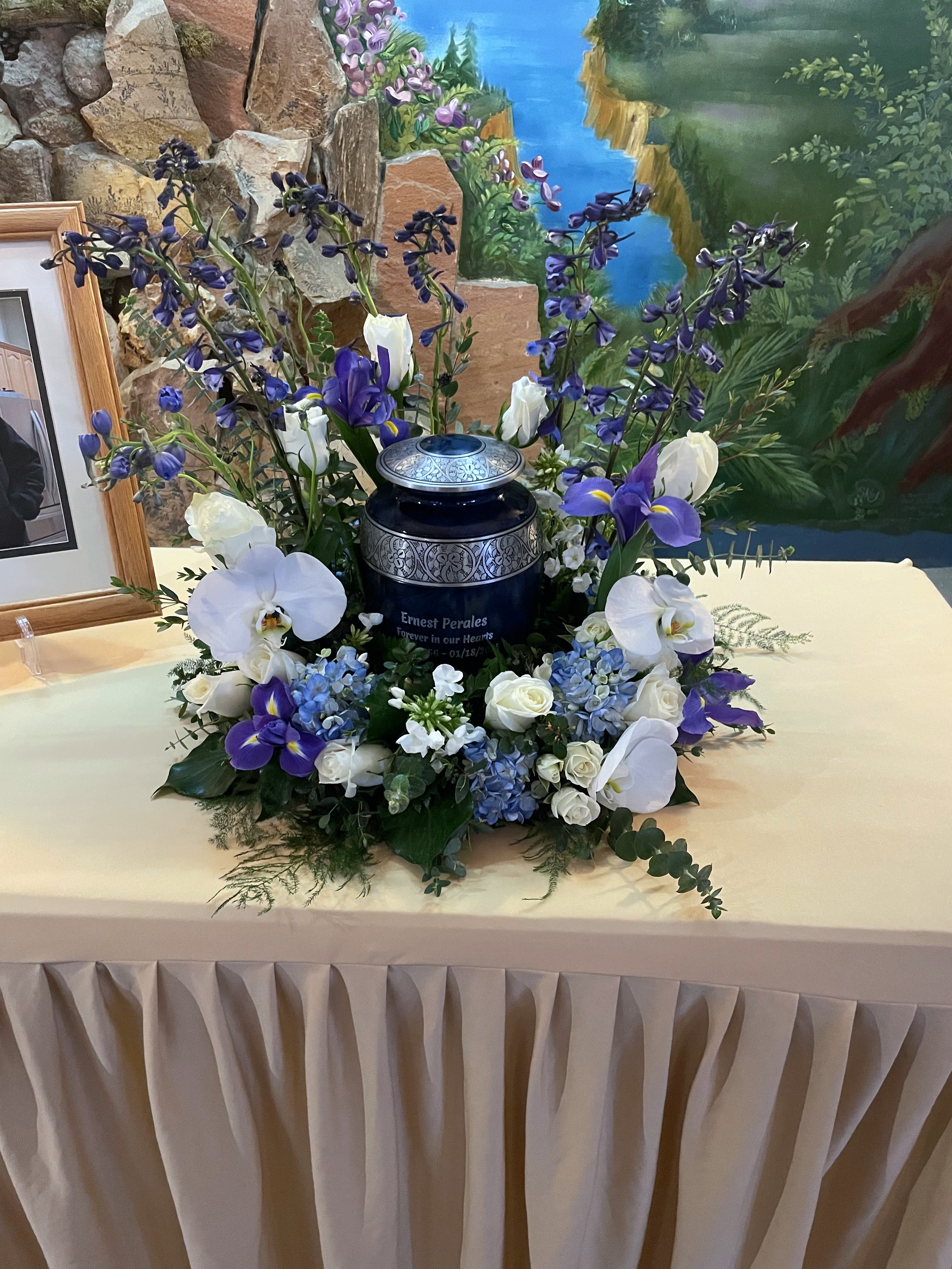 Cremation urn surrounded by blue hydrangeas, white roses, orchids and purple flowers on a draped table