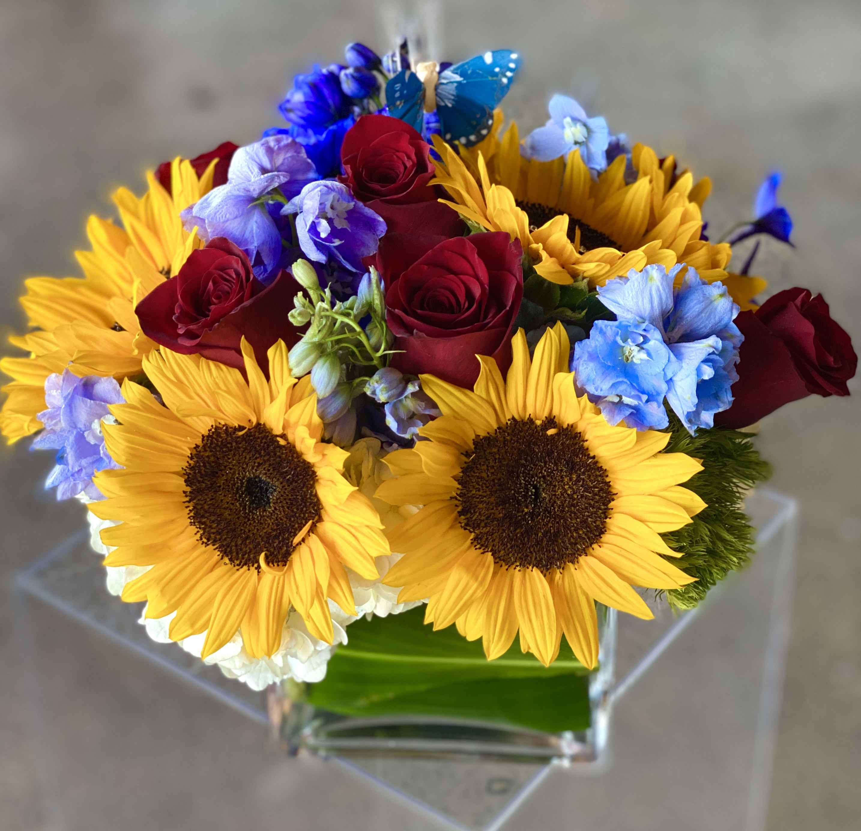 Bouquet of sunflowers, red roses, and blue flowers in a clear vase
