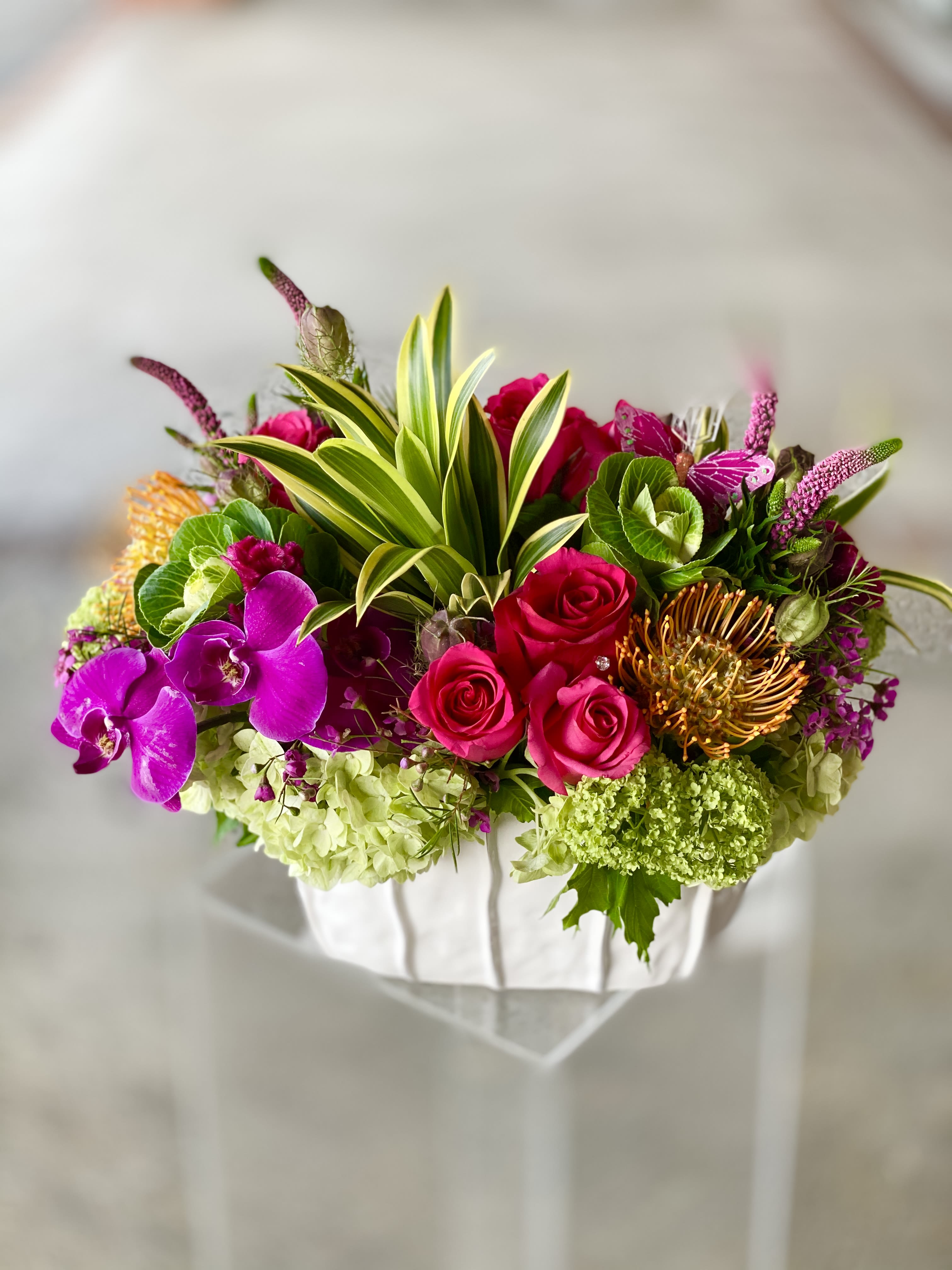 Mookie - Tropical, Colorful flowers Are a Home Run - A tropical abundance full of vibrant hues made of roses, orchids, floral cabbage a lot more, make this a for sure home run. Modern, youthful and fun this is a beautiful design in a textured ceramic vase. 
