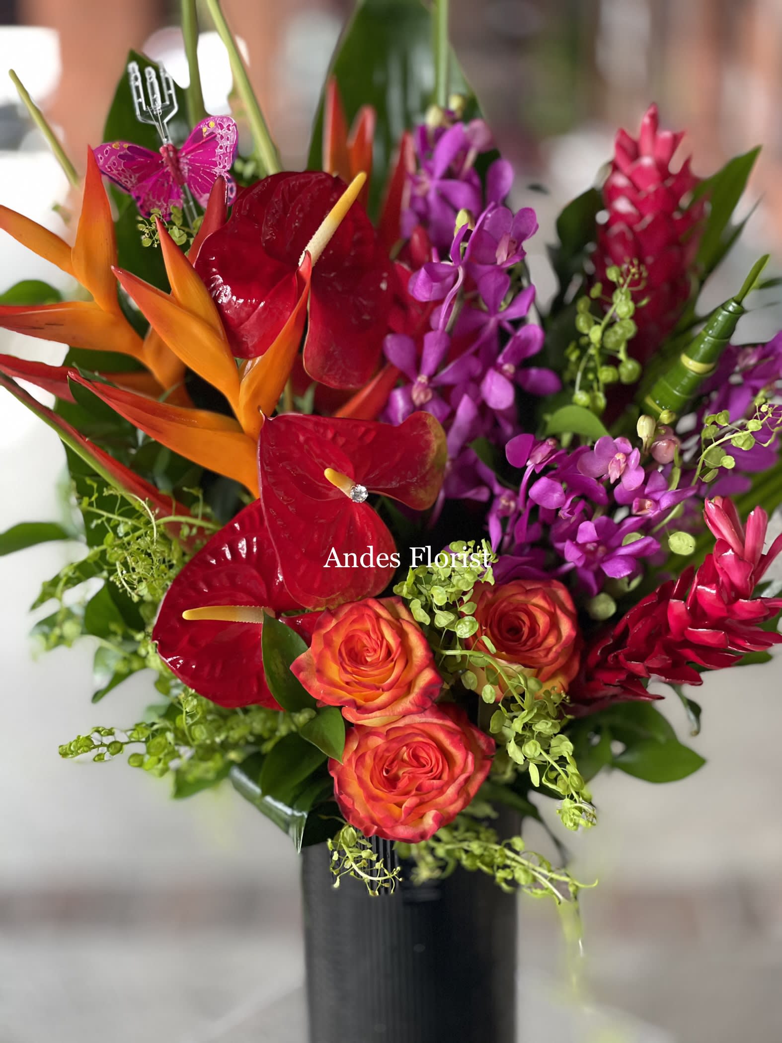 Tropical bouquet with red anthuriums, orange heliconia, and purple orchids in a black vase