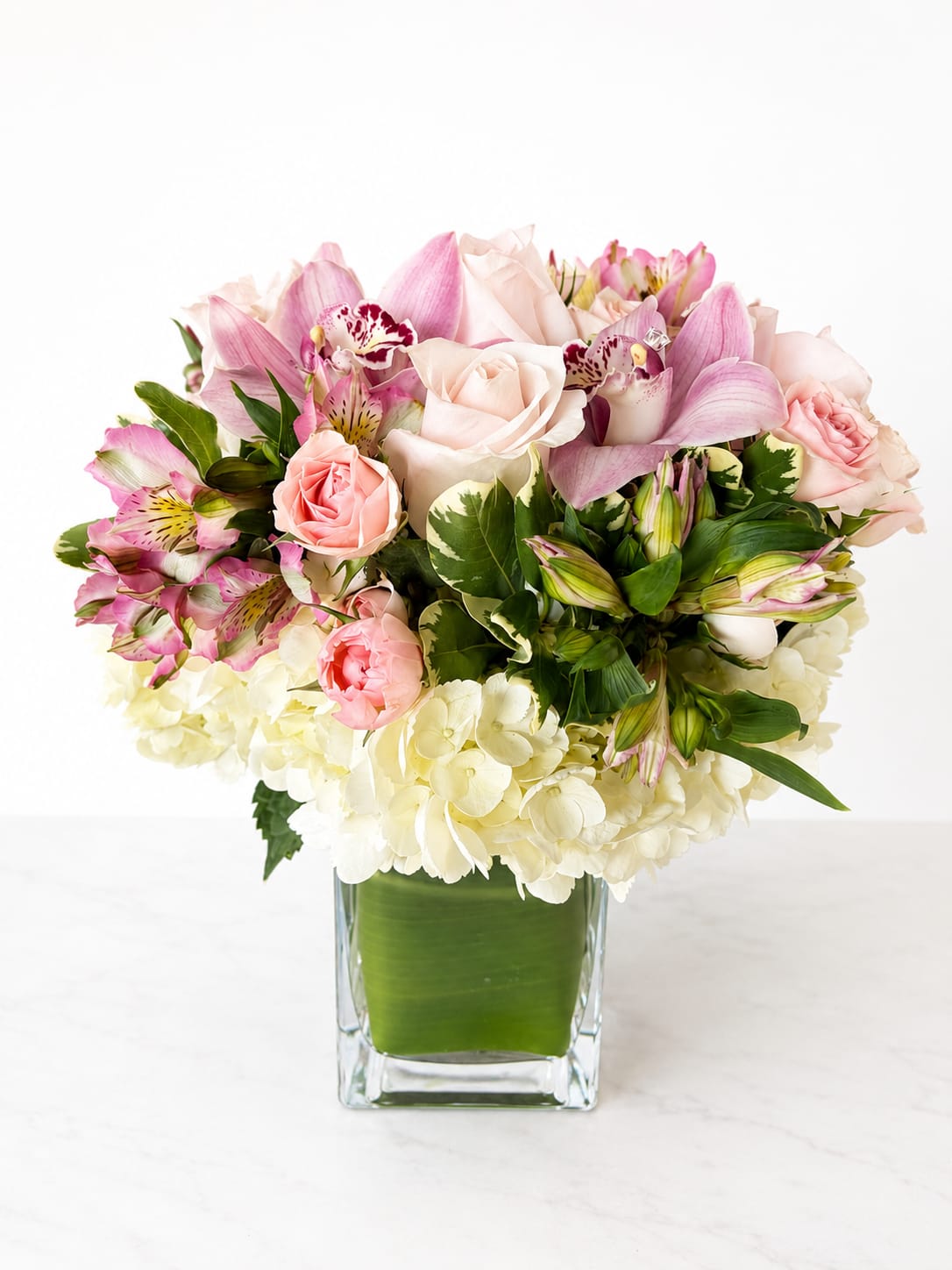 The Darla Blush  - A graceful and romantic arrangement designed in soft blush and ivory tones, featuring elegant cymbidium orchids, delicate garden roses, and fresh seasonal blooms nestled in lush greenery. Designed in a modern glass cube with a clean leaf wrap, this piece offers a perfect balance of sophistication and charm. It’s a timeless expression of love, appreciation, and gentle beauty.