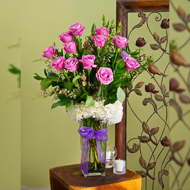 Pink roses in a clear glass vase with white flowers and a purple ribbon