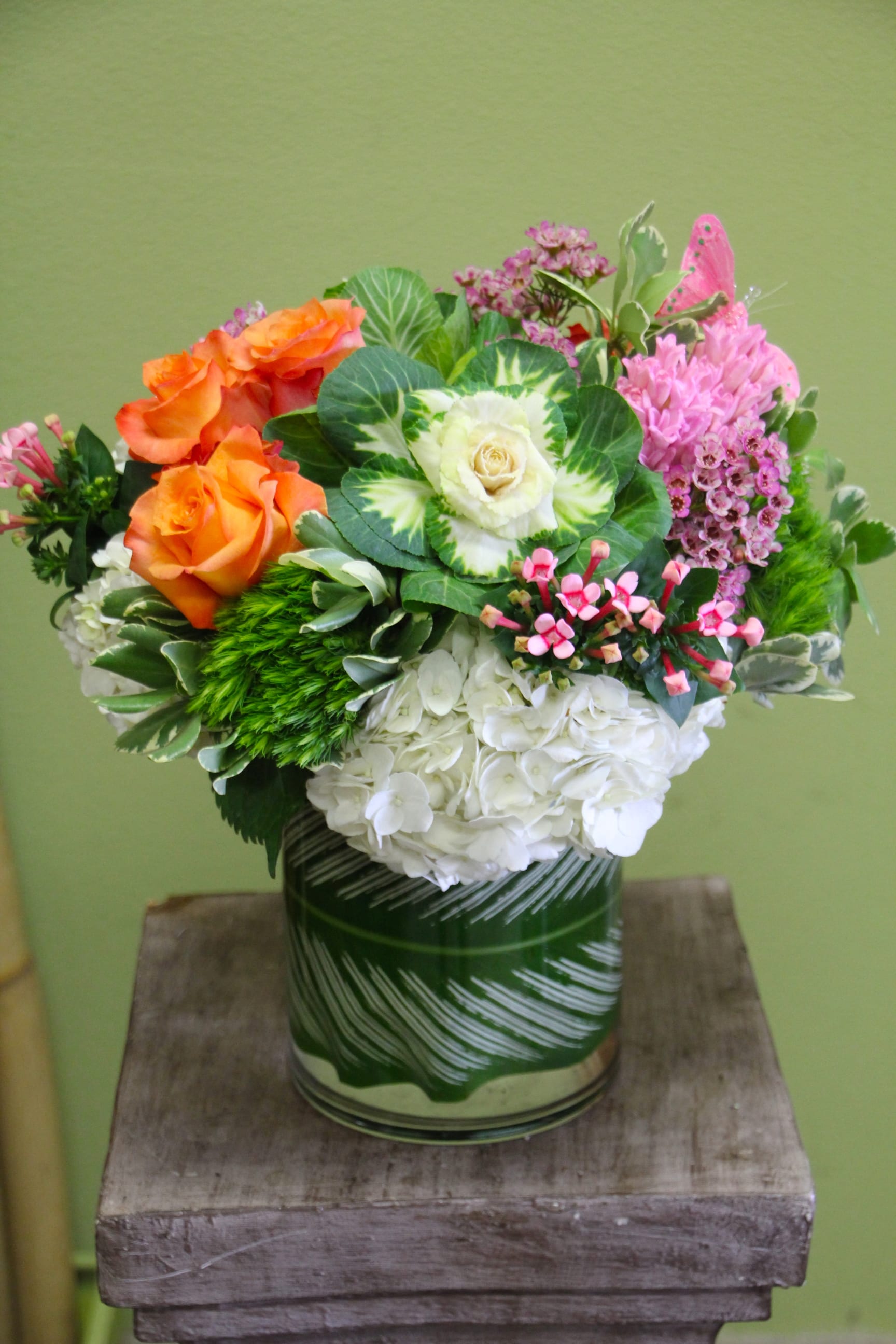 Brenda - Garden Look in a vase! - This abundance of Orange Roses, Kale flowers, Pink Hyacinth, White Hydrangeas, Bavaria flowers, Dianthus flowers, and wax flowers in a glass vase with an exotic leaf will bring your garden into the home!