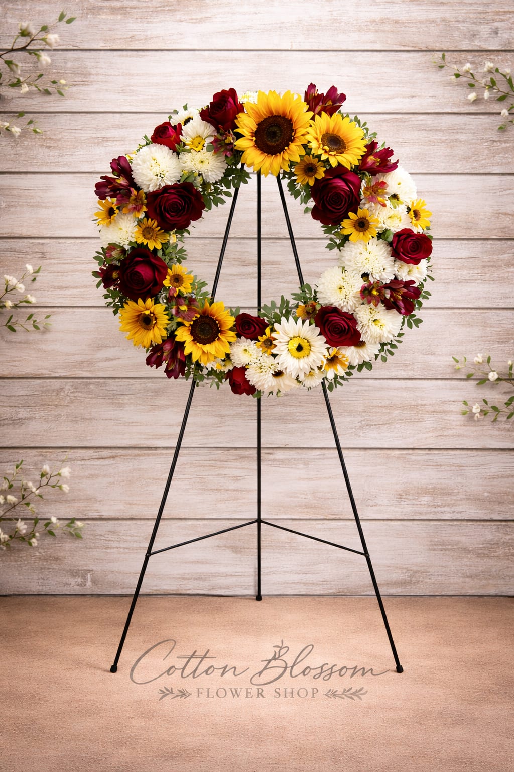 Circular wreath of sunflowers, red roses, and white daisies on a black stand