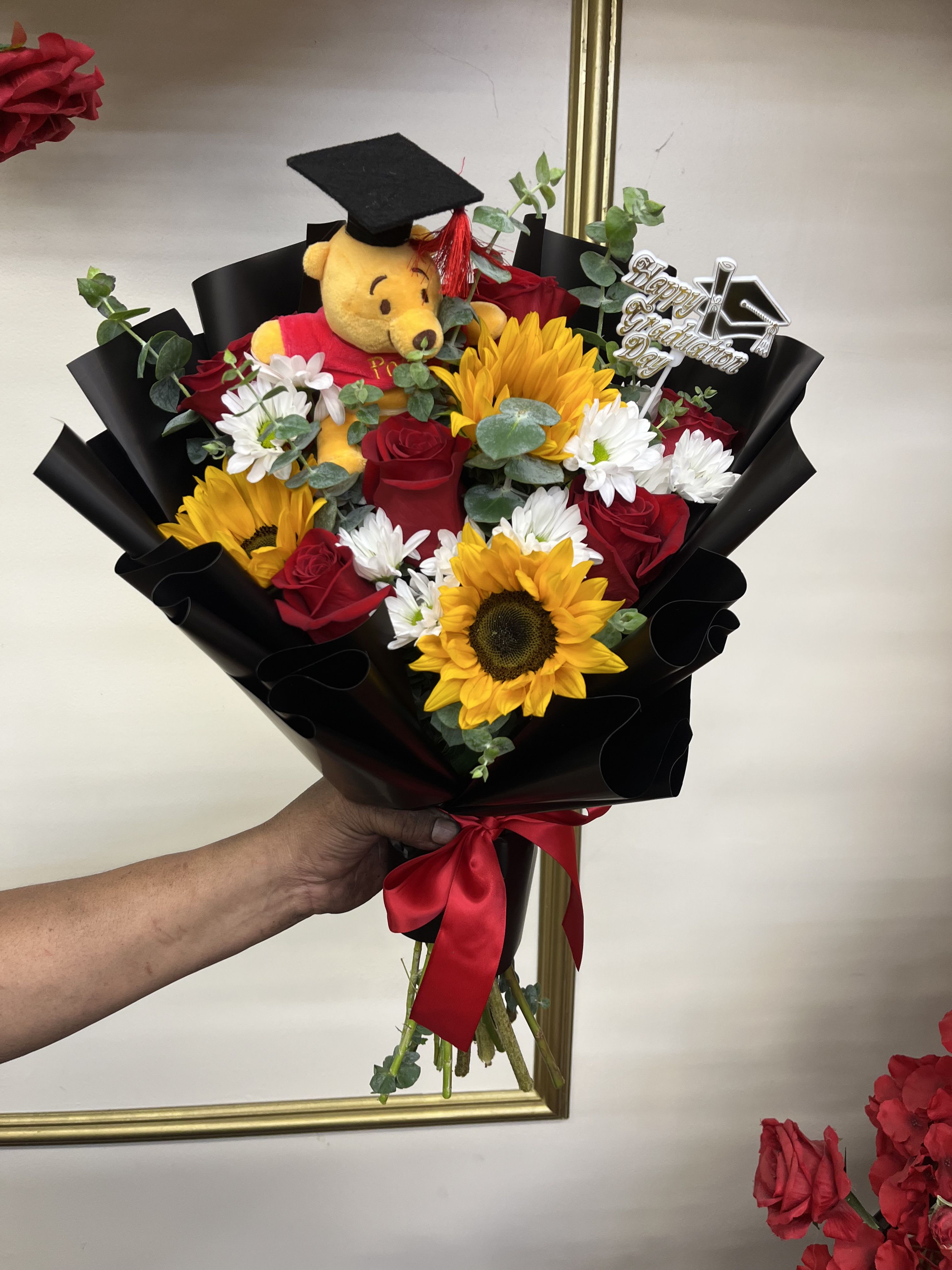 Honey Graduate Bqt - Inspired by everyone’s favorite bear, this graduation bouquet blends warm sunflowers, beautiful roses, and a graduation Pooh plush for the perfect mix of cute and celebratory. A sweet gift for someone starting their next adventure.