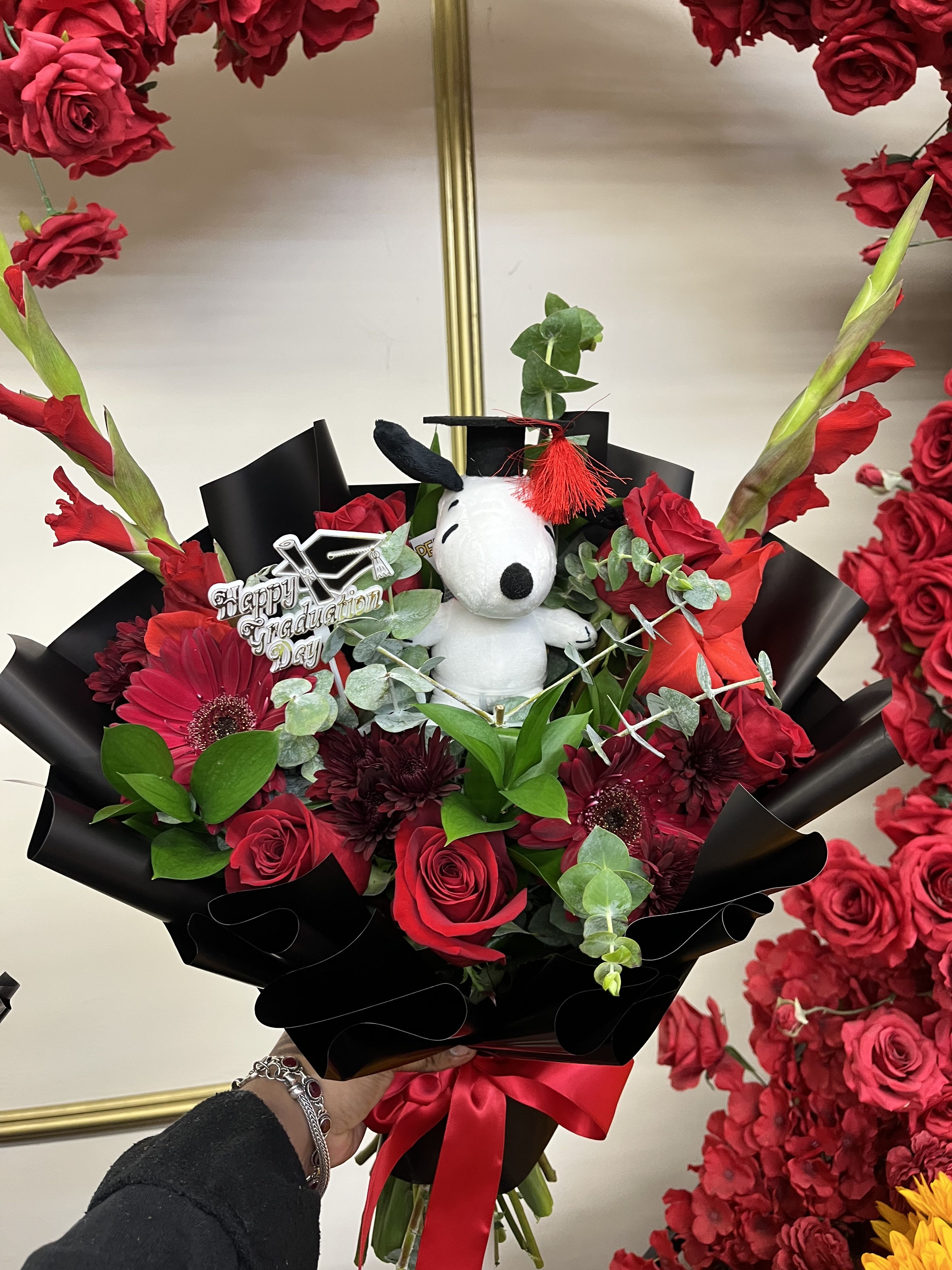 Snoopy Grad bqt - Bright, bold, and full of celebration! This graduation bouquet combines elegant red roses, towering gladiolus, playful gerberas, and pom poms paired with a Snoopy graduation plush for the perfect keepsake gift to congratulate your favorite grad.