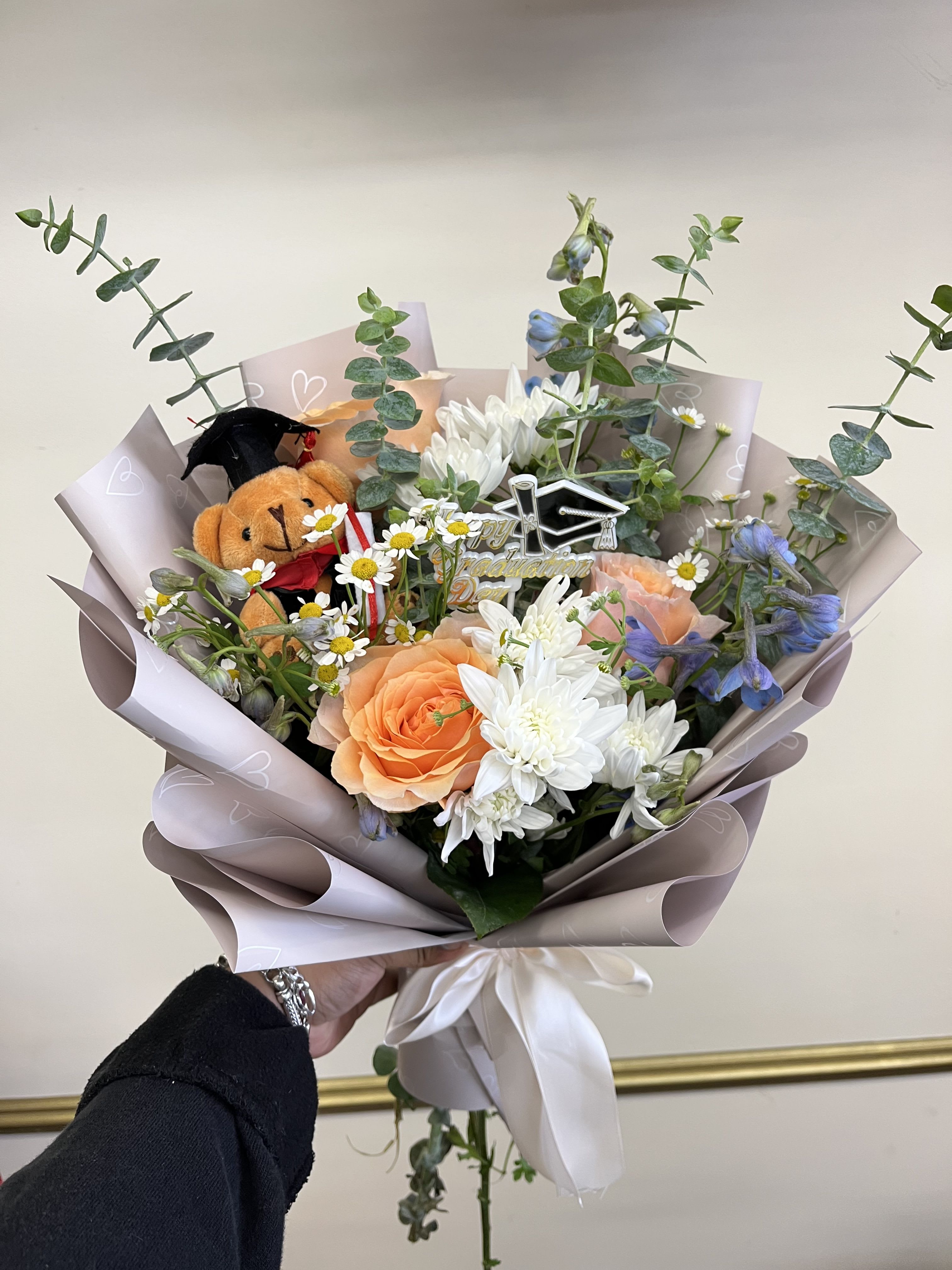 Skybound Success Bqt - Celebrate their achievement with a beautifully crafted graduation bouquet featuring soft peach roses, delicate white pom daisies, striking blue delphinium, fresh eucalyptus, and an adorable graduation bear plush. A timeless arrangement symbolizing growth, success, and bright futures ahead.