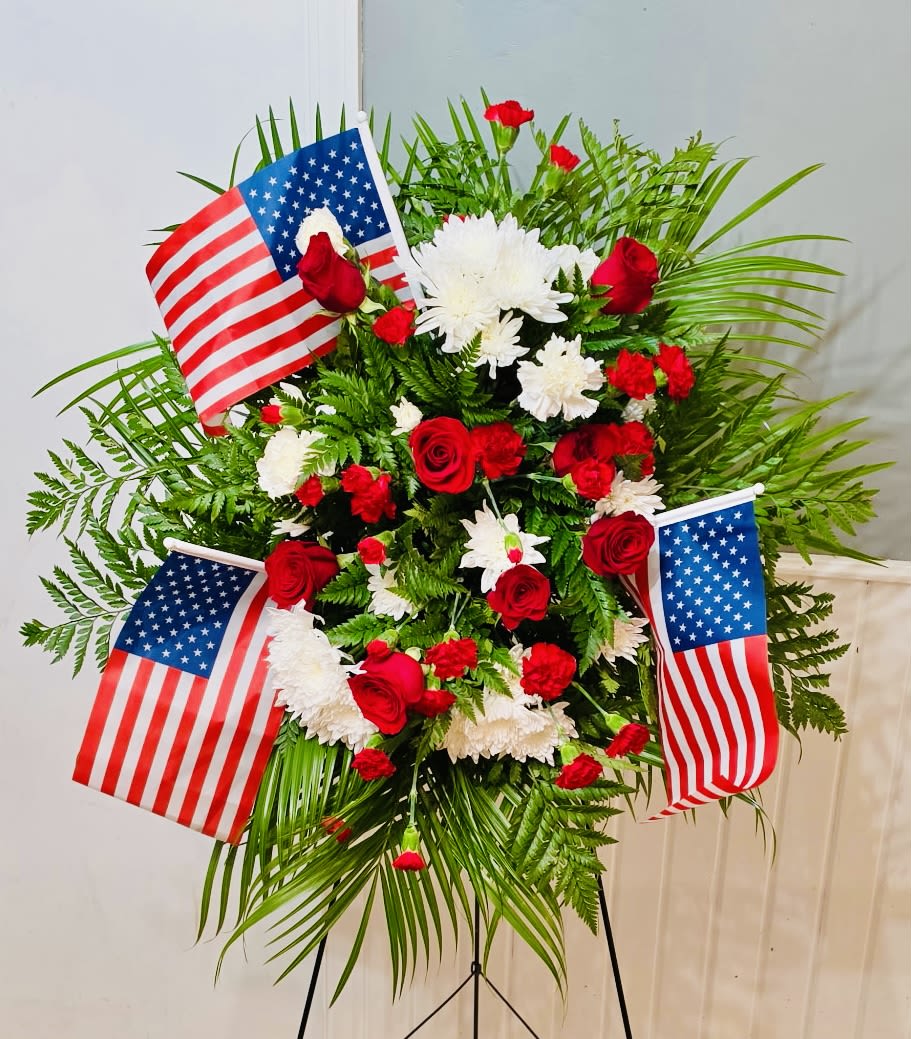 Patriotic Tribute Spray - A formal standing spray composed of deep red roses, red carnations, and pure white chrysanthemums creates a respectful, patriotic tribute-perfect for Memorial Day, Veterans Day, or a funeral honoring service. The arrangement reads as a dignified ceremonial piece; large American flags are placed throughout the design so the person you're remembering is honored with clear national symbolism and heartfelt color.  Dense, layered blooms form an upright, full composition with abundant greenery framing the flowers and adding height and texture. The mix of velvety roses, ruffled carnations, and rounded chrysanthemum blooms gives visual depth and a classic funerary silhouette that photographs and displays well beside a casket or at a graveside. A standing easel supports the spray for stable presentation at any observance.  Choose this arrangement when you want the recipient of the tribute to feel publicly remembered and respected-an unmistakable sign of honor for a loved one who served or for patriotic commemorations. Standard sizing and care are set for ceremonial use; the easel and flags are included so it arrives ready to display.
