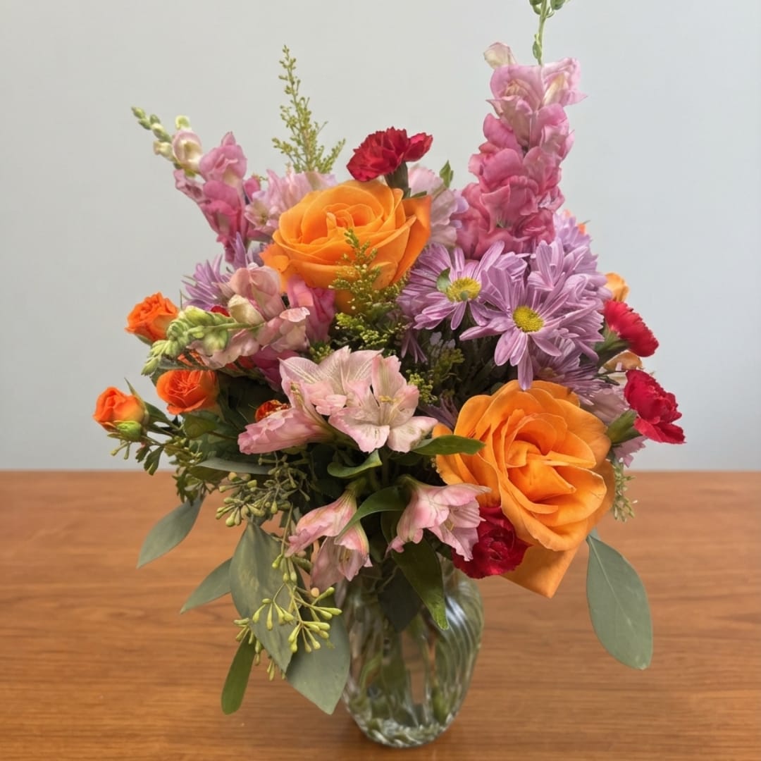 Mixed bouquet of orange roses, pink snapdragons, and purple daisies in a glass vase
