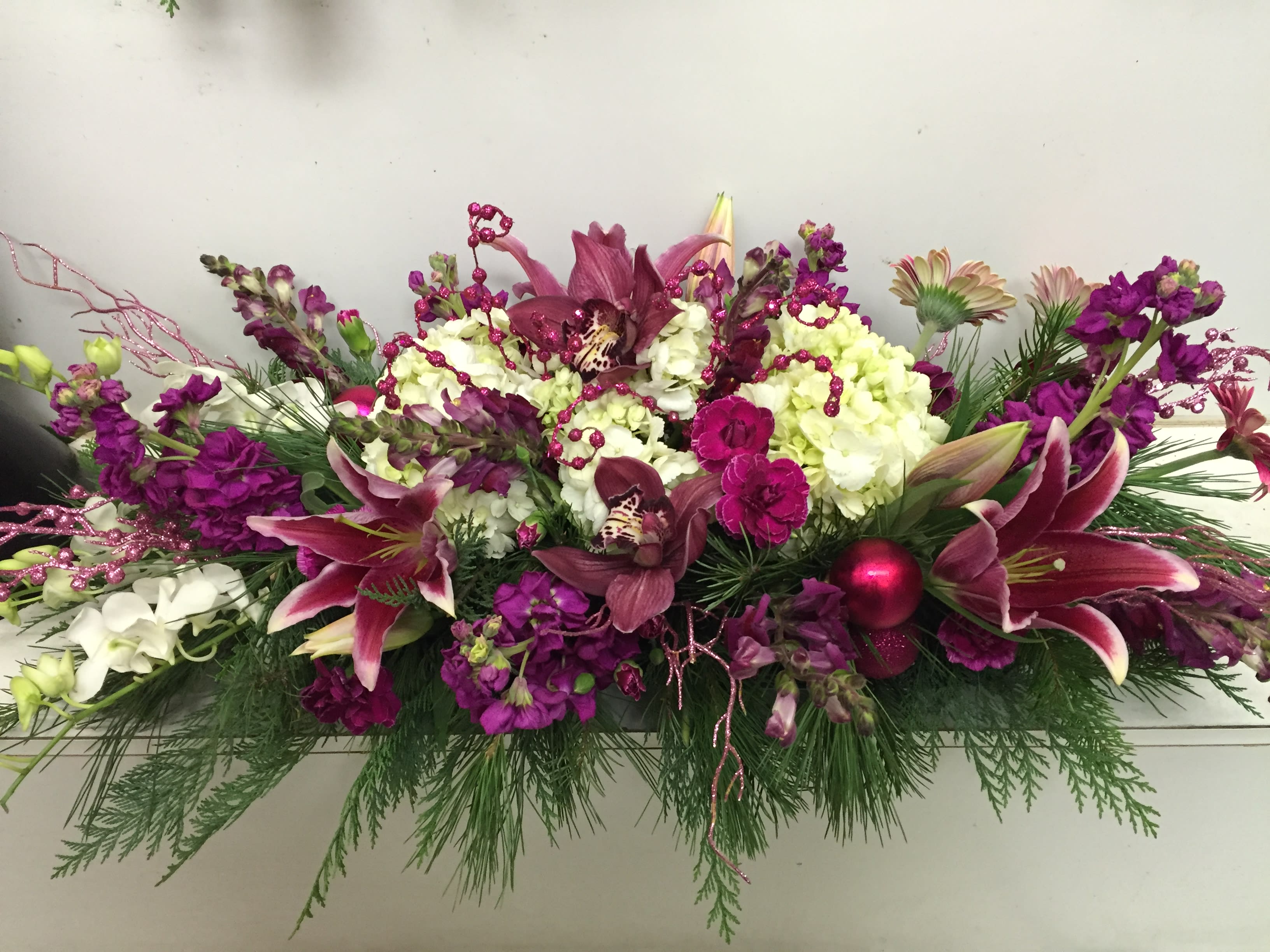Christmas Burgundy Centerpiece - Star Gazers, Ivory Hyrdrageas, Burgundy Stock, Snapdragons, White Cymbidium Orchids, Dendrobium Orchid, Gerbia Daisies, Christmas Greens with Christmas ornaments, pearls, and branches. 25" long Dimensions: Low Plastic Container (not shown) 20" long Seasonal: all seasonal flowers, christmas greens available christmas only Substitute Available for any colors or occasion. For example, all white funeral piece.   