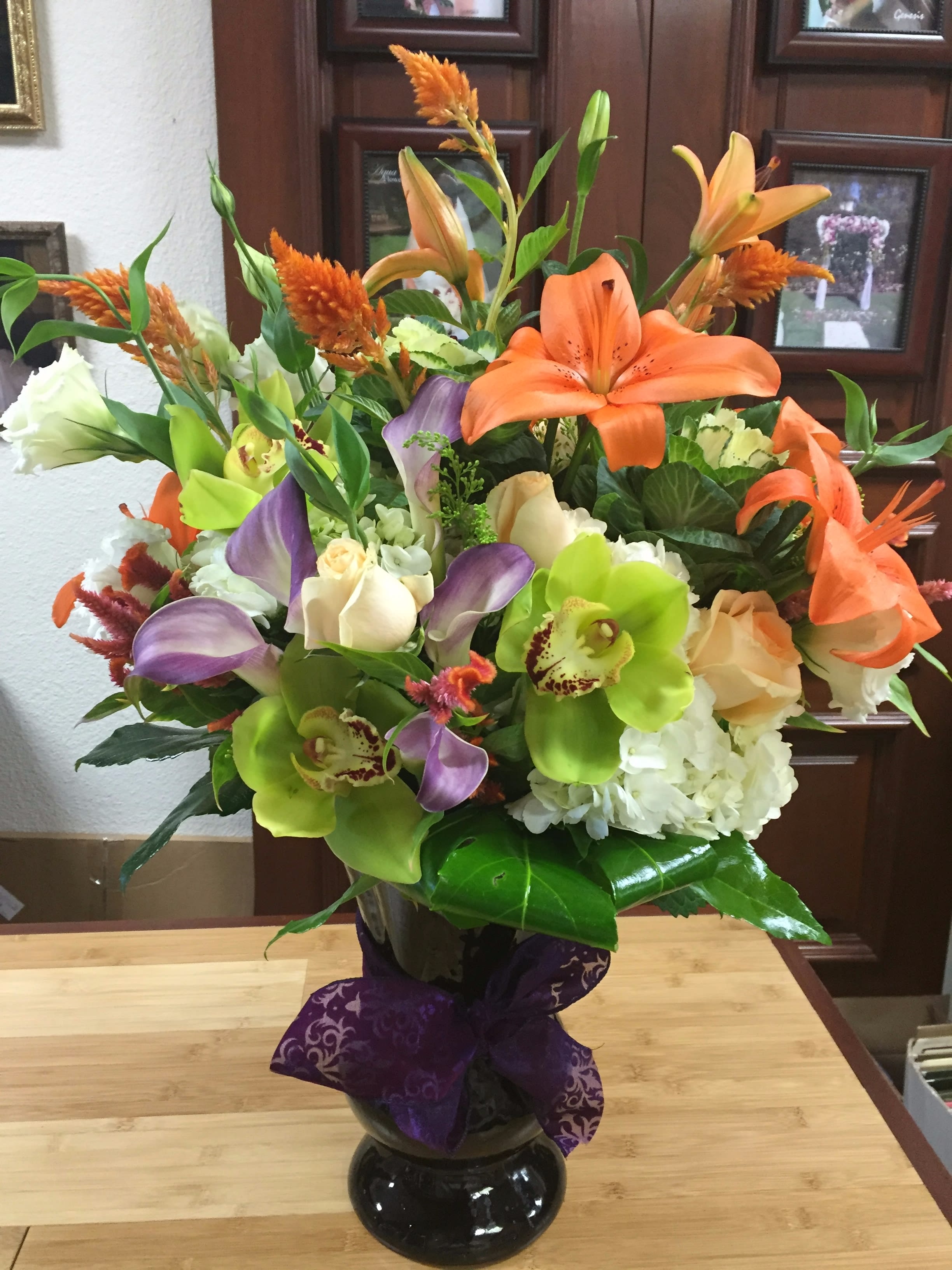 October Vibes - Type of Flowers: Orange Lilies. Mini-purple Calla Lilies, Orange and white roses, ivory hydrangeas, White Lisianthus, Orange Celosia, Green Cymbidium Orchids, Cabbage in a purple vase with purple ribbon Dimensions: Purple vase  Seasonal Flowers:Yes Substitute Available in different colors or flowers. 
