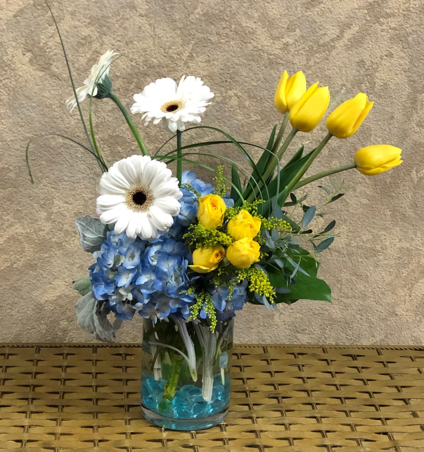 Glow and Grow - Type of Flowers: Blue Hydrangeas, White Gerbera Daisies, Yellow Tulips, Yellow Spray Roses and greens in a short vase with blue crystals. Availability: All year round Substitute Available: Yes Design View: Front Facing Facing View Photo shown: Regular 