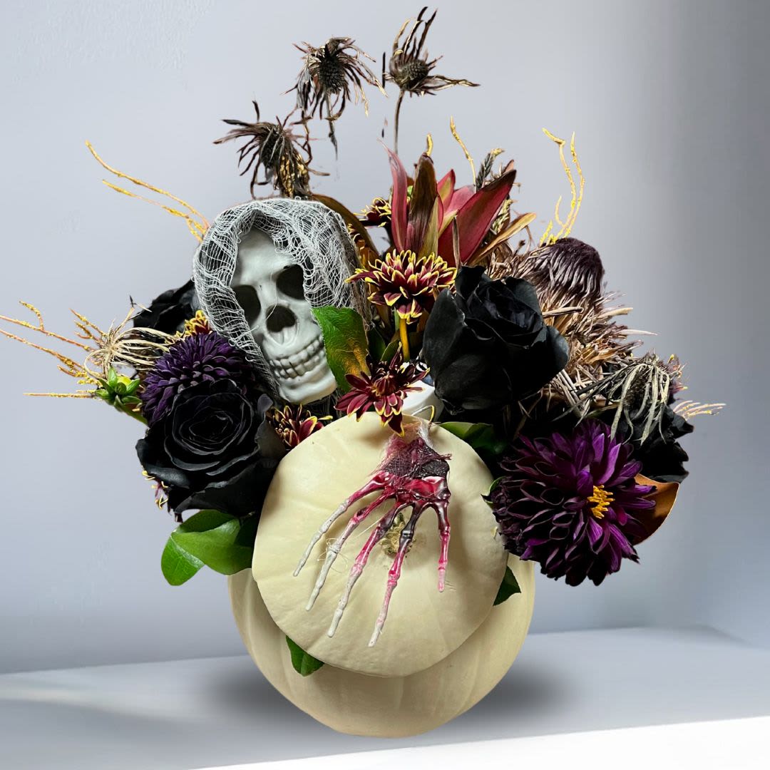 Dark and Moody Jack O'latern Pumpkin - Have fun this spooky speason with this halloween aesthetic! Recipe: Dark Roses, Purple Dahlias, Red Chrysanthemums, Safari, Dried flowers, and more with a skeleton face and crawling bloody hand in a Natural White Pumpkin and light! Availability: October Substitute Available: Yes Design View: Asymmetric Front Facing View Photo shown: Regular