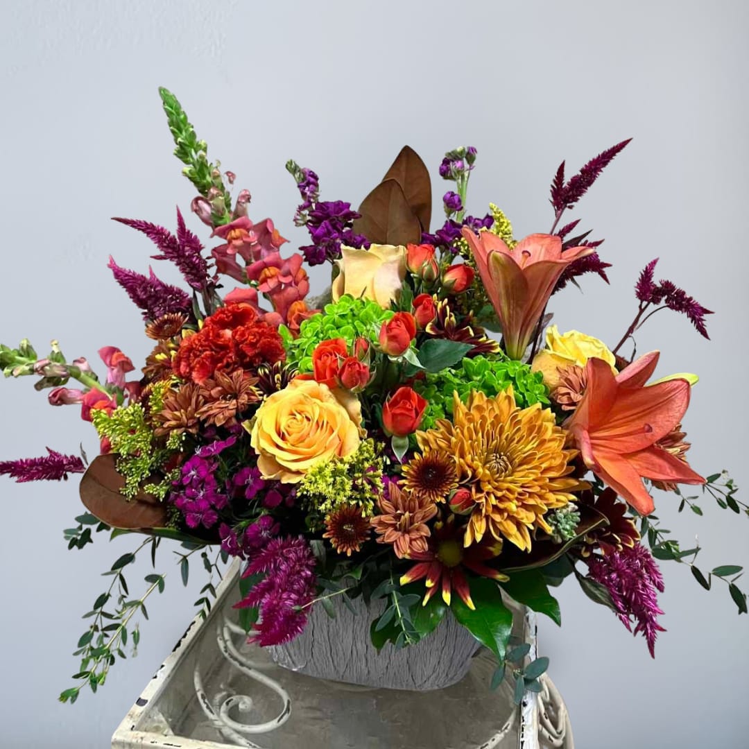 Stay Humble - Recipe: Brown Chrysanthemums, Orange Roses, Spray Roses, Blood Orange Lilies, Snapdragons, Protea, Green Hydrangeas, Fuchsia Amaranthus, Stock, Daisies, Eucalyptus and Magnolia Leaves in a white oval, ceramic vase. Availability: All year round Design View: Symmetric Front Facing View Photo shown: Standard
