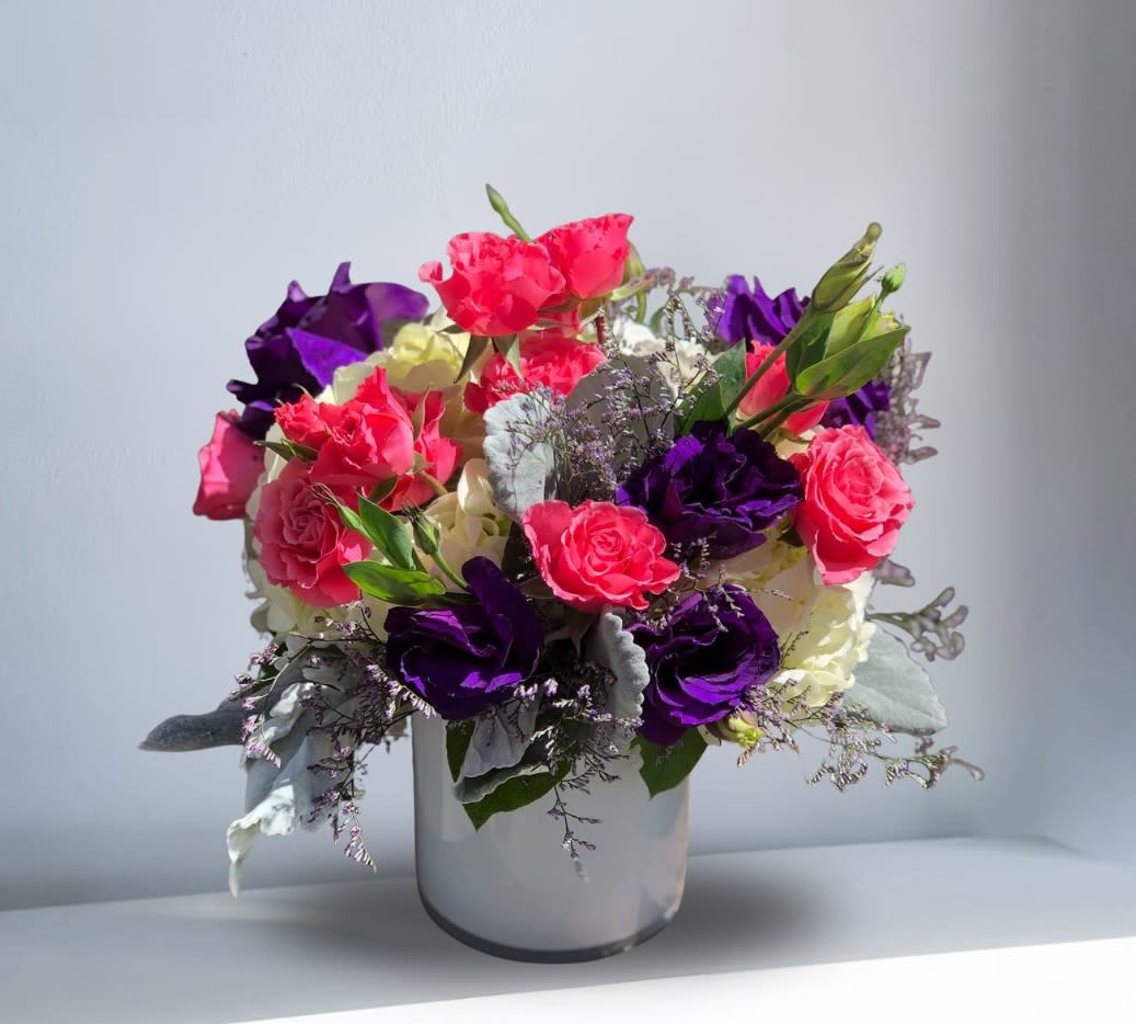 Color Crush - Turn heads with this striking, modern arrangement! Deep, velvety purples meet vibrant hot pink roses in a lush, eye-catching composition. Perfect for making a statement, celebrating a special occasion, or sending a message that’s as bold and unforgettable as the colors themselves. Recipe: Purple Lisianthus, Hot Pink Roses, White Hydrangeas, Dusty Miller, and Misty Blue in a short, white, cylinder vase. Seasonal: All year round Orientation: All Facing View Photo shown: Standard
