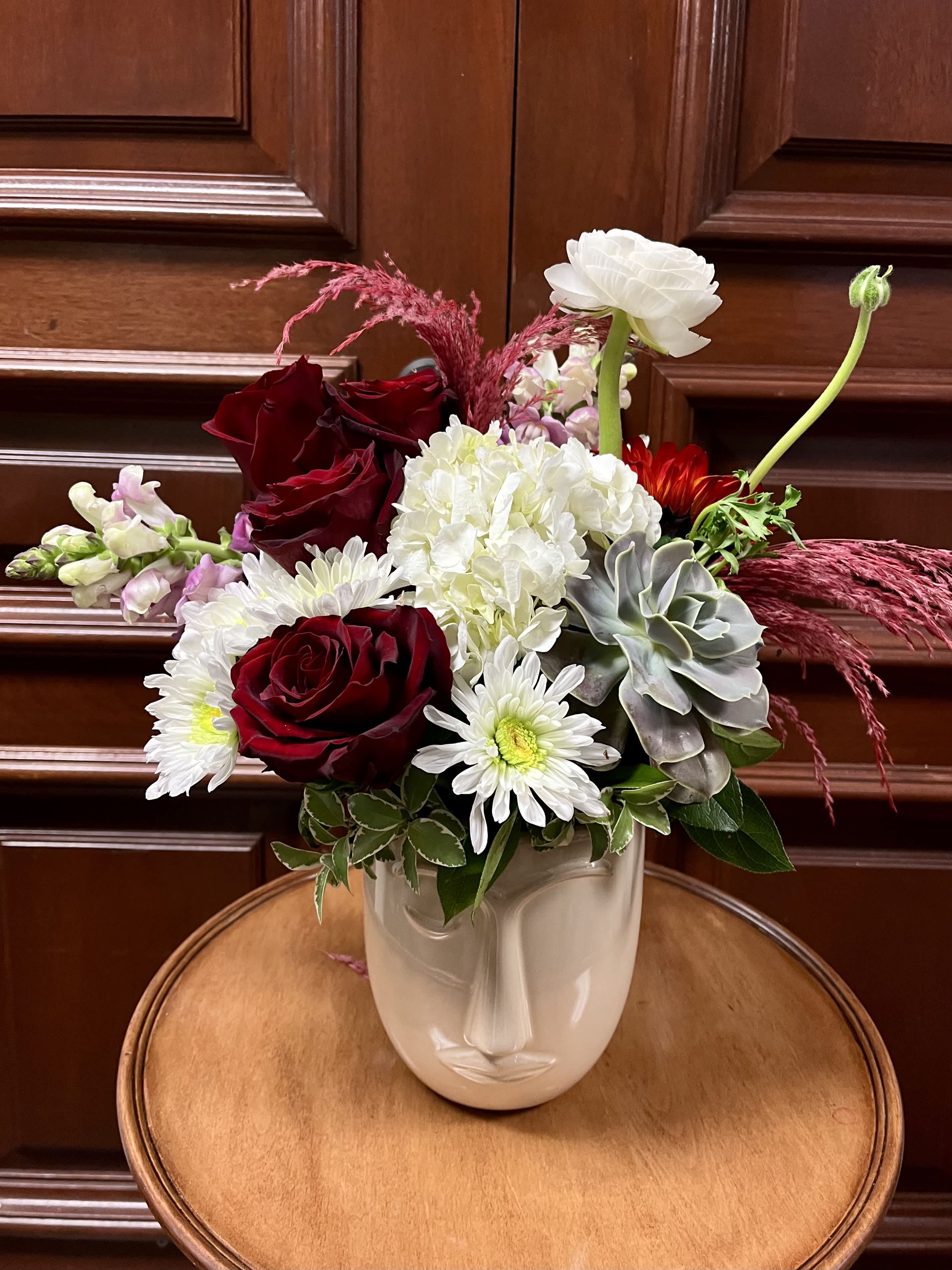 Rosalia NEW - Recipe: White Ranuculus, White Chrysthemums, Pink Snapdragon, Red Roses, Sunflower, and Succulent, with Pink Feather Accents in a face vase. Availability: All year round Design View: Symmetric Front Facing View Photo shown: Standard 