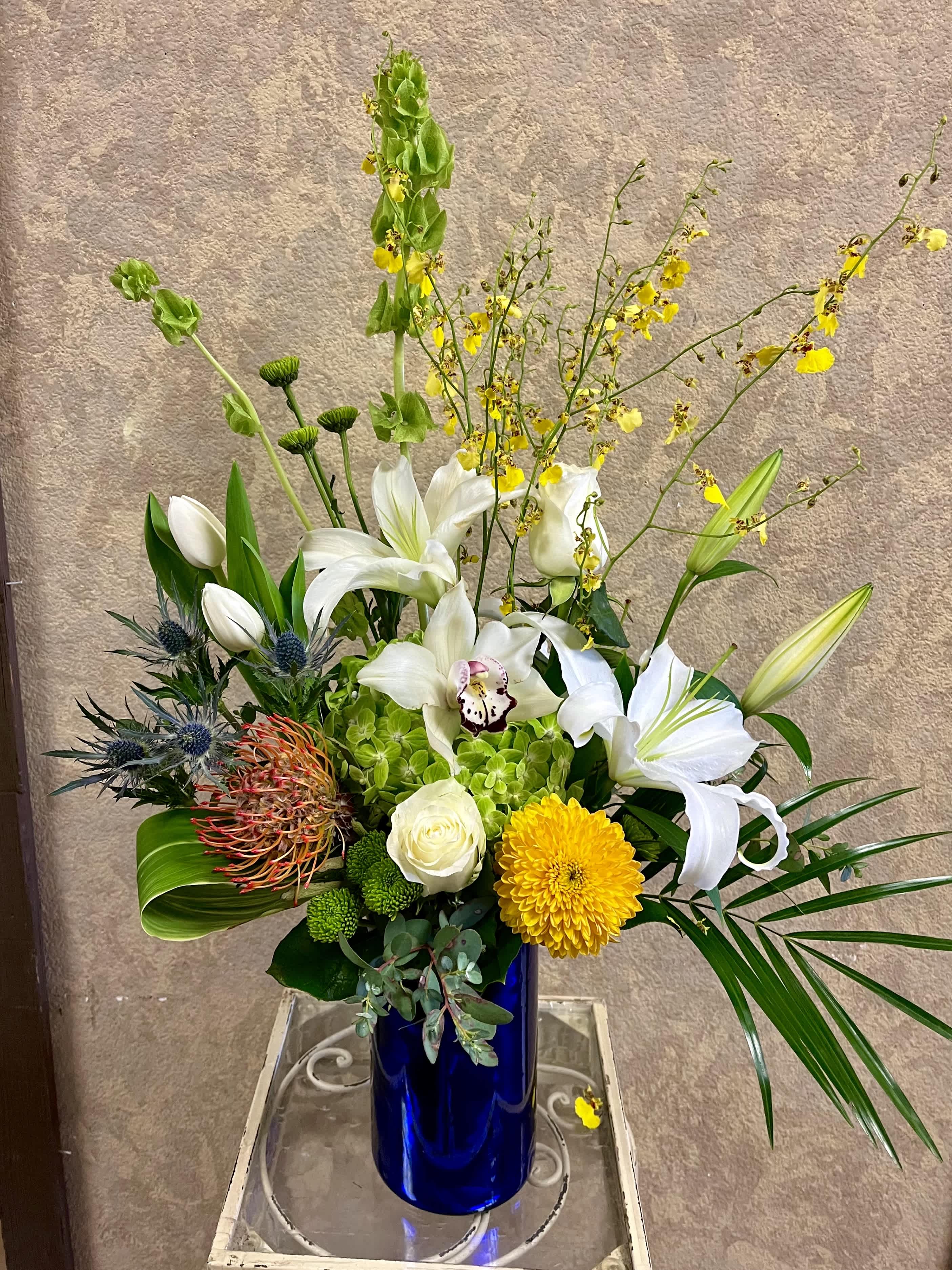Okayama, Japan - Recipe: White Casablanca lilies, White Tulips, Yellow Tropical Oncidium Orchids, Bells of Ireland, Protea, Green Hydrangae, White Roses, White Cymbidium Orchid, Eryngium and greens in a tall, blue vase. Season: Seasonal Tropical Oncidium Orchids Orientation: Front Facing View Photo shown: Standard 