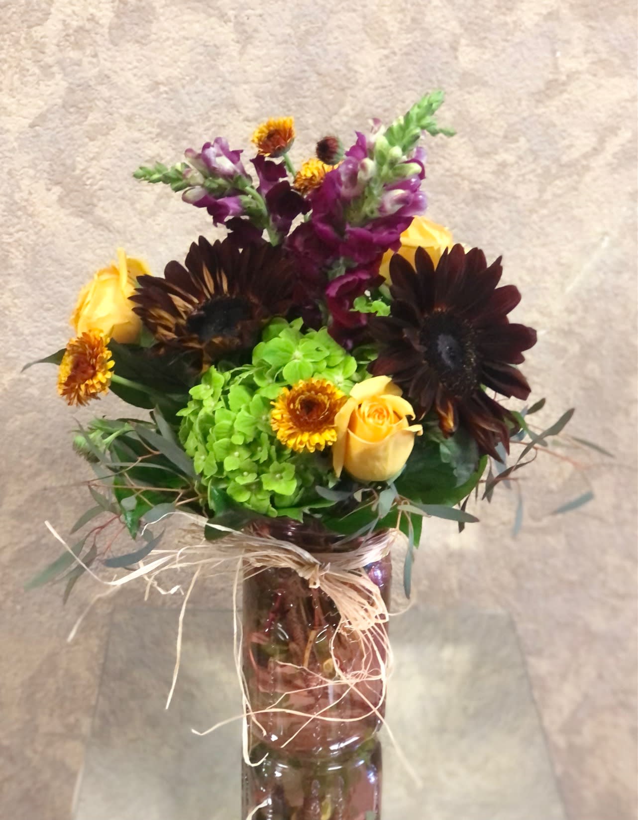 Morticia - Type of Flowers: Dark Sunflowers, Yellow Roses, Green Hydrangea, Yellow Daisies, Purple Snapdragons in an orange mason jar vase wrapped with straw ribbon. Design View: All Facing View Photo shown: Standard 