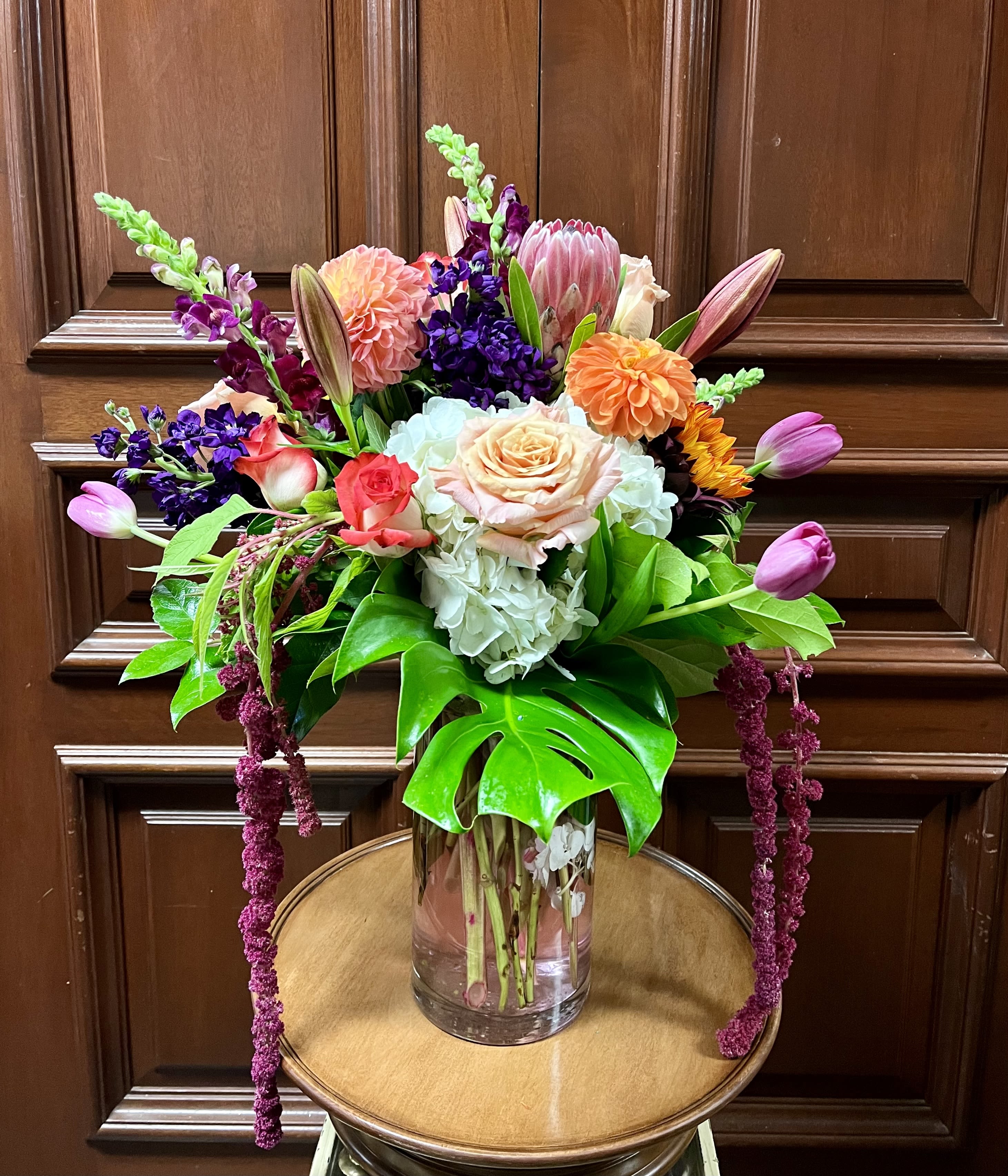 Mariella - Recipe: Yellow Seasonal Dahlias Red/Orange Lilies, Red/Orange Sunflower, Pink Snapdragons, Orange Roses, PInk Roses, Protea, White Hydrangae, Orange Tulips, Purple Stock, Green Amaranthus and Orange Carnations in a tall, crystal clear vase. Availability: Seasonal Dahlias Design View: Symmetric Front Facing View Photo shown: Standard 