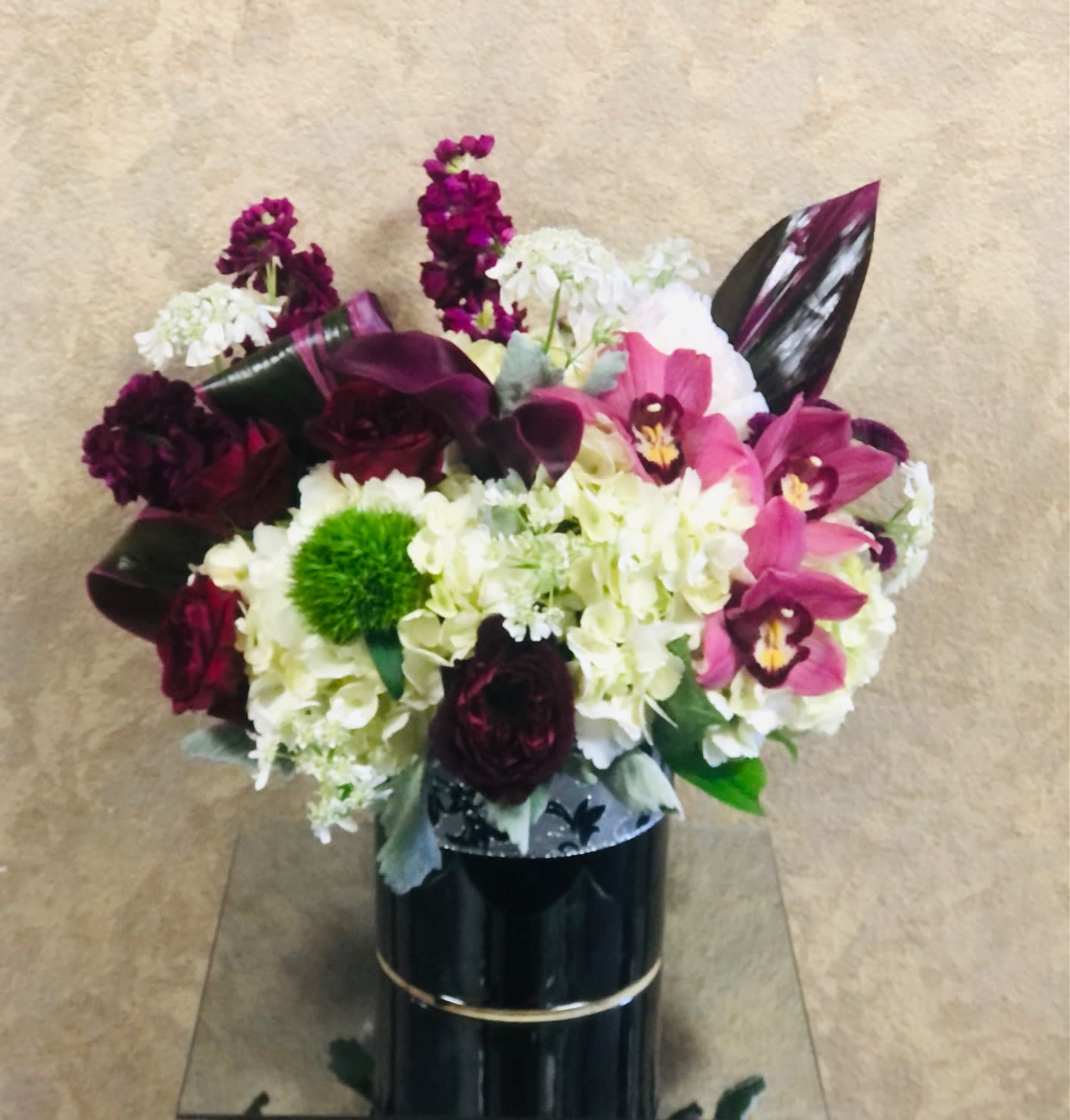 Genavi - mini calla lilies, burgundy, stock, burgundy roses, orchids, and hydrangeas in a black vase with a silver ribbon. 