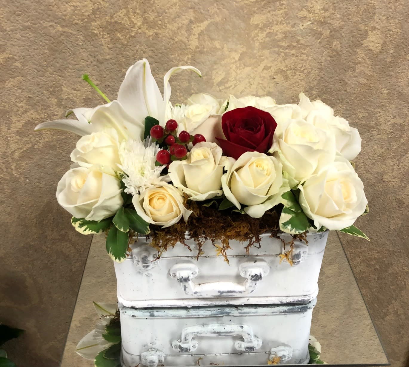 Love Travels Far Distances - Type of Flowers: Asiatic White Lilies, White Roses, Hypericum Berries, and Red rose in a white, plaster suitcase. Availability: All year round Design View: Symmetric Front Facing View Photo shown: Regular 