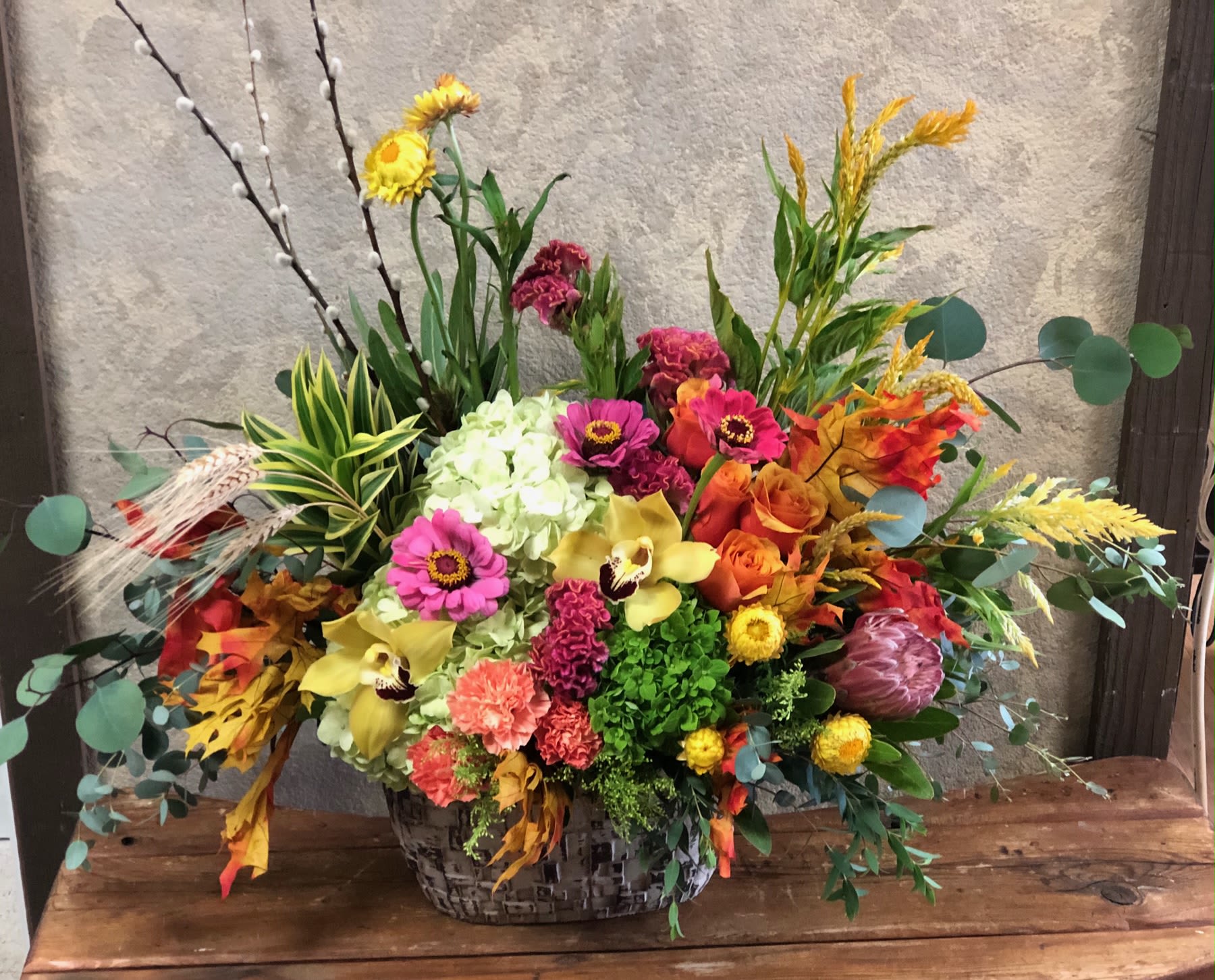 Summer Meets Autumn - Recipe: Stunning bright, fall arrangement including Zinnias, Green Hydrangeas, Orange Roses, Yellow, Orchids, Strawflowers, Protea and more in a plaster, wood like design vase. Availability: All year round Orientation: Symmetric Front Facing View Photo shown: Standard