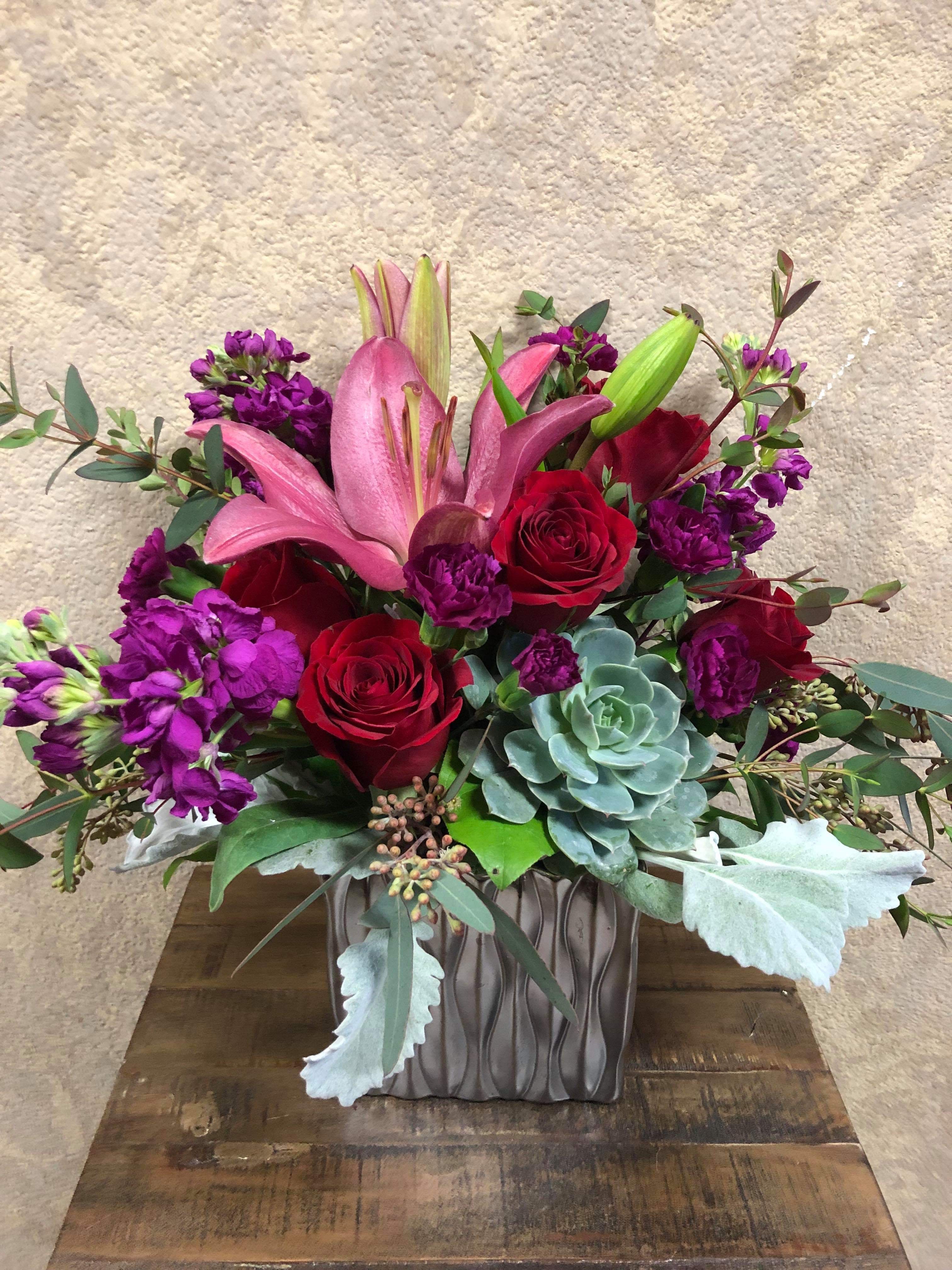 A Winter's Love - This romantic design has a mix of dark purples, passionate reds, and dusty blues, she is going to love! Recipe: Red Roses, Purple Stock, Pink Asiatic Lilies, Succulent and Dusty Miller in a 6" silver, cubed vase. Seasonal: All year round Substition: Silver vase is no longer available. Will sub for similiar Orientation: Front Facing View Photo Shown: Standard
