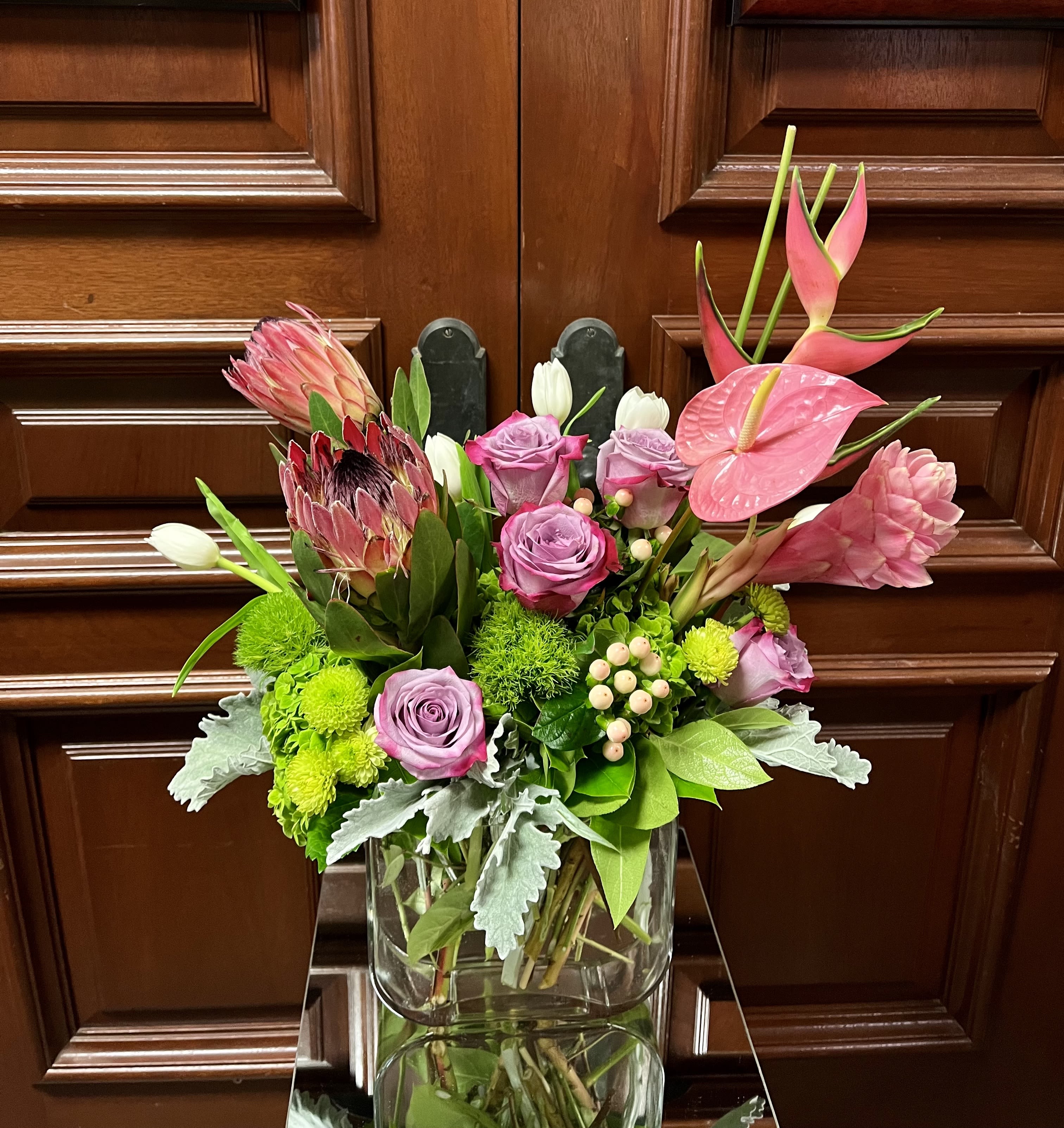 Paradise is Calling - Recipe: Ginger, Tropical Protea, Birds of Paradise, Anthurium, Lavender Roses, White Tulips, Hypericum Berries, Dianthus Barbatus, Green Buttons, and Dusty Miller in a clear, square, skinny vase. Availability: All year round Design View: Symmetric Front Facing View Photo shown: Standard