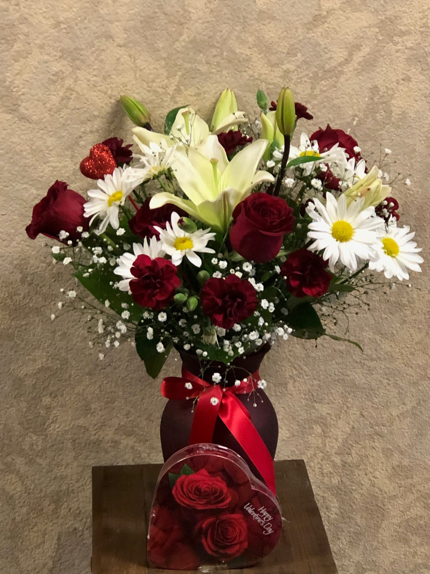 The Love Package - Type of Flowers: Traditional Valentine's Day Design with White Asiatic Lillies, Red Roses, Red  Carnations and White Daisies in a tall red vase with heart stick and red ribbon. Availability: All year round Substitute Available: Yes Photo shown: Premium Chocolates available for purchase separately.