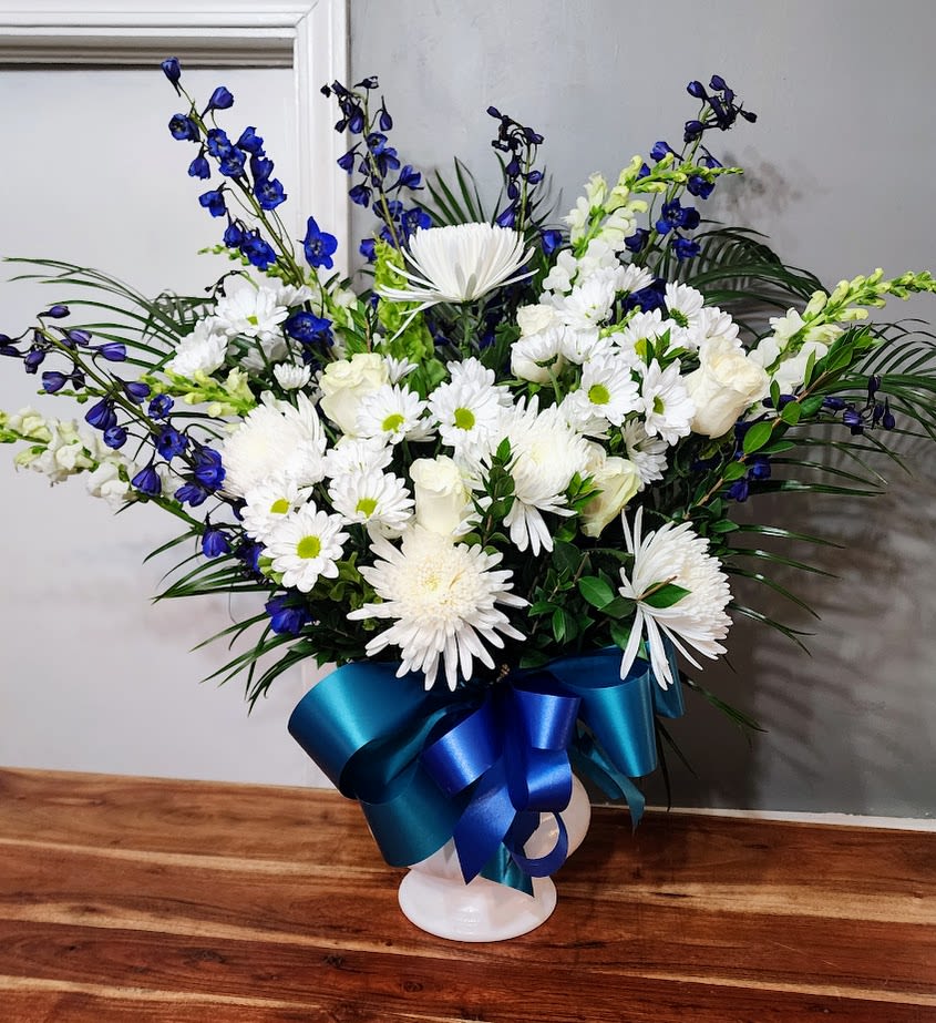 Royal Glory - A fan-shaped spray of crisp white chrysanthemums and daisies, softened by white roses and snapdragons with tall blue delphinium spires creates a quietly majestic sympathy arrangement that comforts at a difficult time. As a sympathy and funeral tribute or a Thinking of You gift, Royal Glory says calm, steady support-the person you're thinking of will feel seen and soothed by its composed, serene presence.  This arrangement is presented in a rounded white pedestal vase and finished with a glossy blue satin ribbon bow tied low at the base, adding a formal, polished touch. The palette of white, blue, green and mixed color accents keeps the look clean and dignified while the delphinium provides vertical movement and depth. The fan-shaped design reads beautifully on a memorial table or as a bedside pick-me-up; dimensions and exact stem counts are preserved in our store details.  Ideal for winter remembrances, Memorial Day displays, or to let someone know you're holding them close, Royal Glory works equally well for a service or a private delivery to express sympathy and steady care. Delivered fresh to West Frankfort, IL, the arrangement arrives ready to display-lightly mist the blooms and change the water every other day to keep the flowers looking their best.