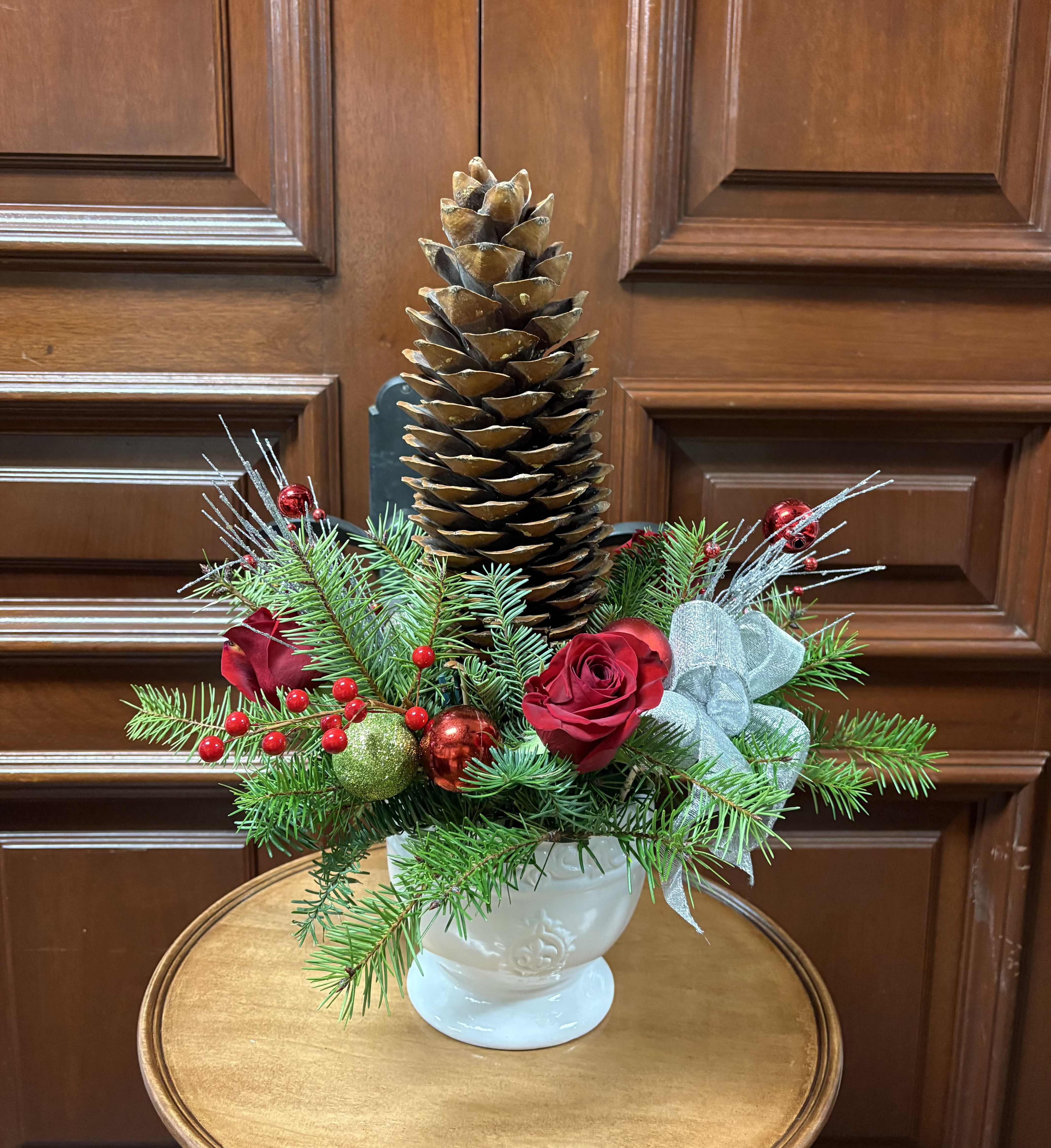The Great Pine cone treasure - This holiday arrangement will last a long time!  Recipe: Red roses, Red faux berries, pine greens, Pine branches, Silver branches, Holiday ornaments and silver ribbon in a white, round ceramic vase with stem. Seasonal: Winter Orientation: All Facing View Photo shown: Standard 