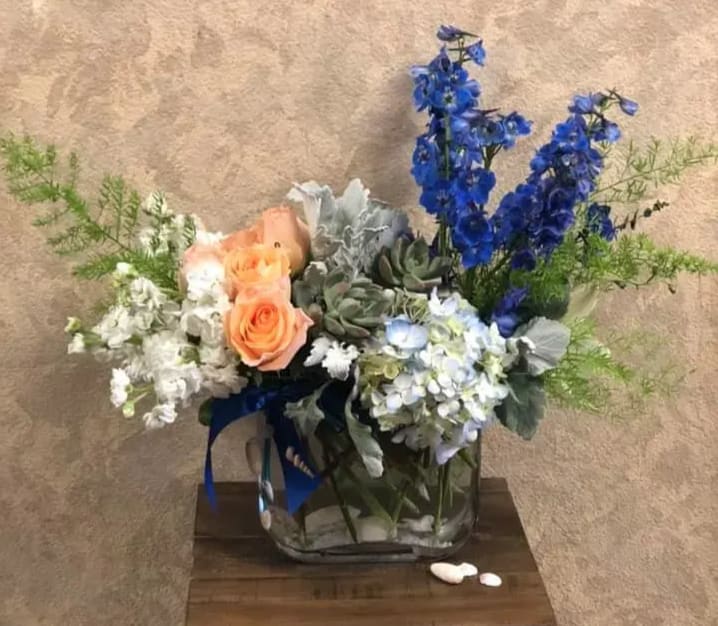 The Sea of Cortez - Where the Sea meets the Desert. The Sea of Cortez, Baja California Sur represents the most beautiful sunsets and clear waters. Recipe: Blue Delphinium, White Stock, Blue Hydrangea, Orange Roses, Duty Miller, Fern, Succulents and Seashells in vase. Availability: All year round Substitute Available: Yes Design View: Symmetric Front Facing View Photo shown: Regular 