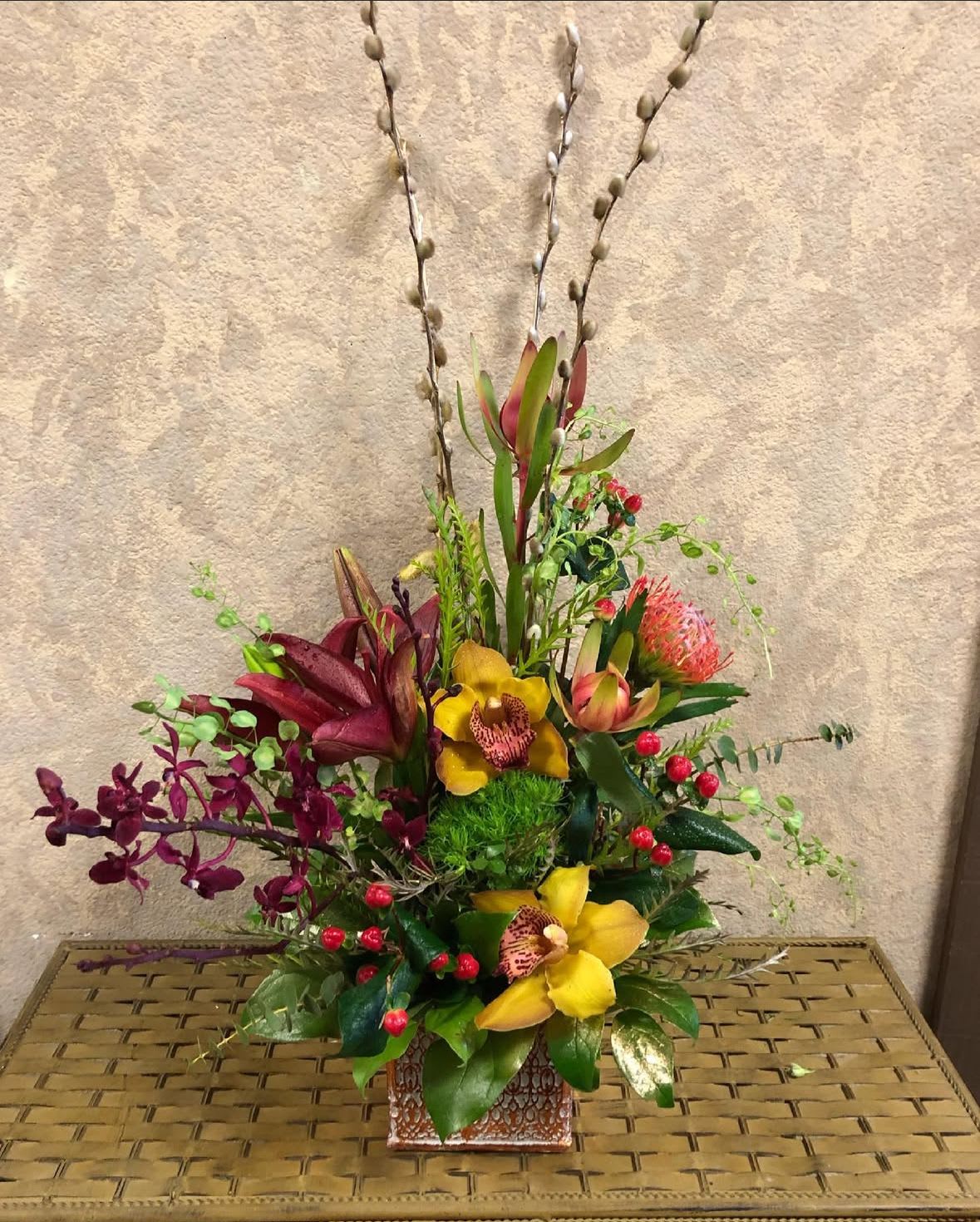 Lunar New Year - Recipe: Red Liliesm, Pin cushion Protea, Red Orchids, Yellow Cymbidium Orchids, White Roses, Safari, Berries, and Pussy Willow in a square, golden vase. Seasonal: All year round Orientation: Front Facing View Photo shown: Standard 