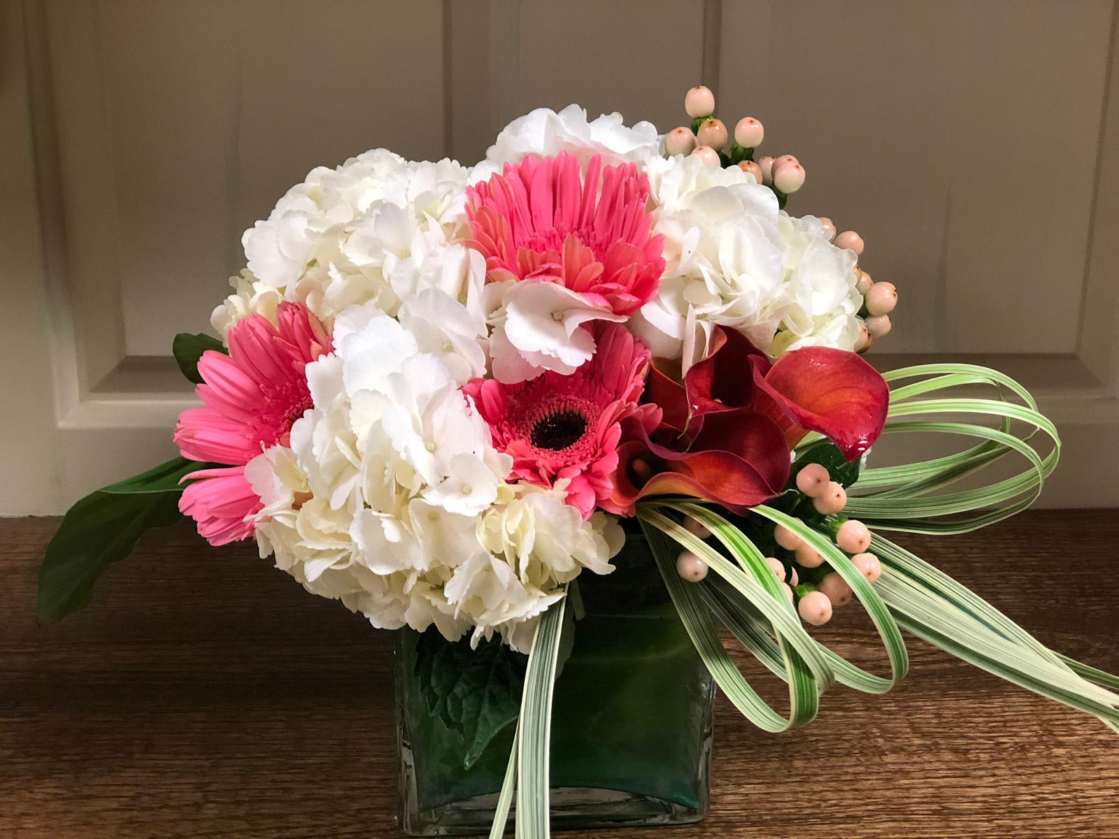 Sweet Baby Girl - Type of Flowers: Gerbera Daisies, Mini Calla Lilies, White Hydrangeas, Hypericum, Aroid Leaves, and Beargrass in a short vase wrapped with a leaf. Availability: All year round Substitute Available: Yes Design View: Symmetric Front Facing View Photo shown: Regular