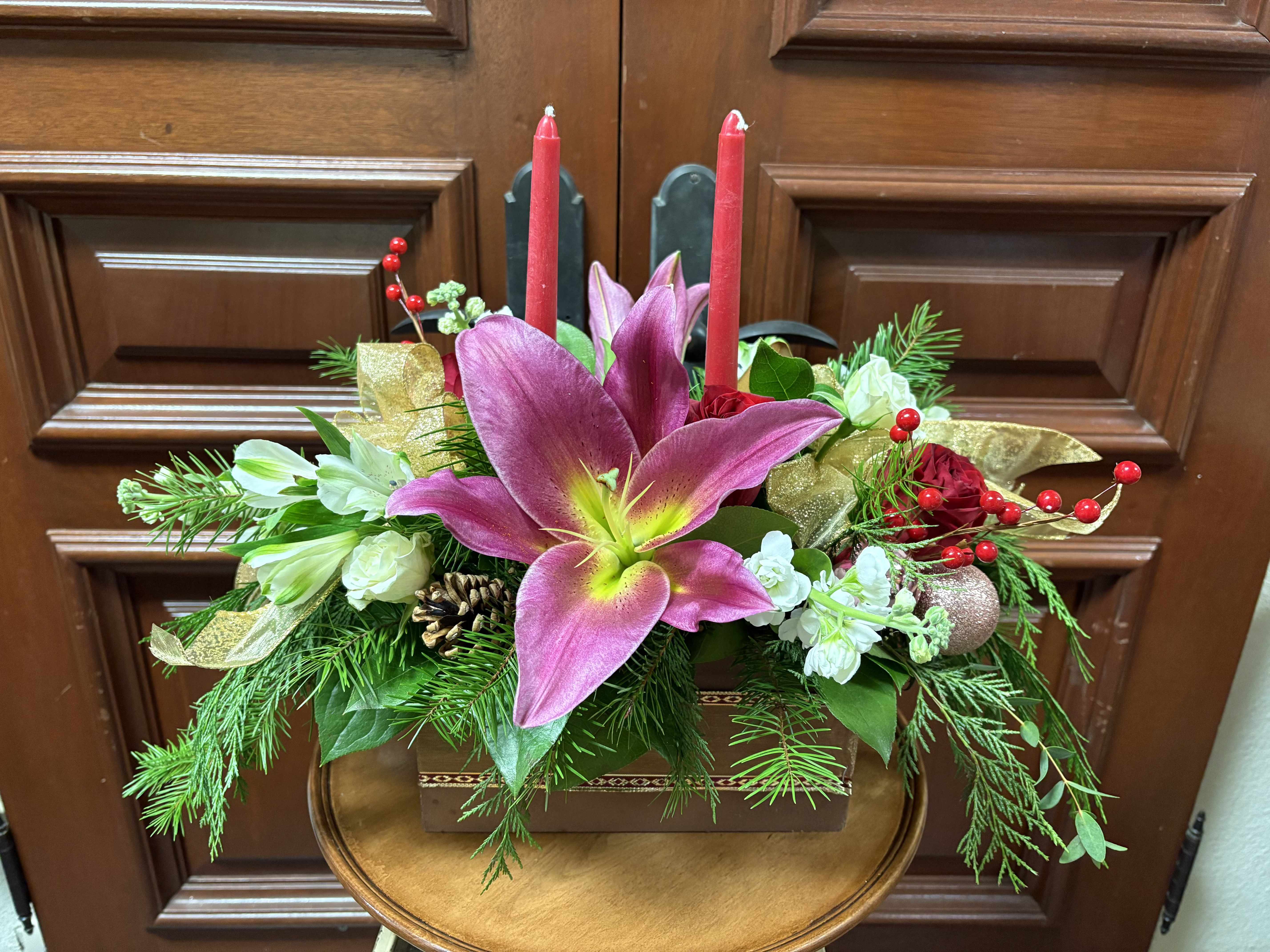 Holiday Cheer Centerpiece - Recipe: Red Roses, Alstromerias, Stock, and Mauve Lilies, Pine Cones, Pine greens, Berries, and rose gold ornaments, with two red candles, and gold ribbon on a wooden box wrapped in ribbon. Availability: Winter Substitute Available: Yes Design View: All Facing View Photo shown: Standar