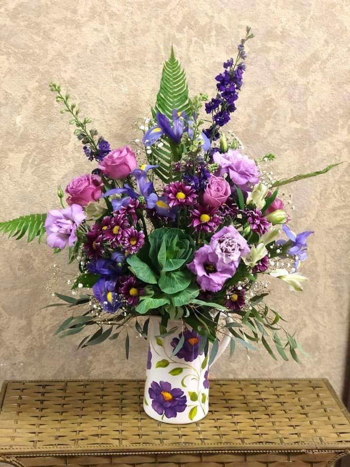 Shades of Purple Garden Pitcher - Type of Flowers: Purple Lackspur, Lavender Roses, Purple Iris, Purple Daisies, Lavender Lisianthus, Green Cabbage, White Alstroemerias, and green succulent in a short vase. Availability: All year round Substitute Available: Yes Design View: Symmetric Front Facing View Photo shown: Regular 