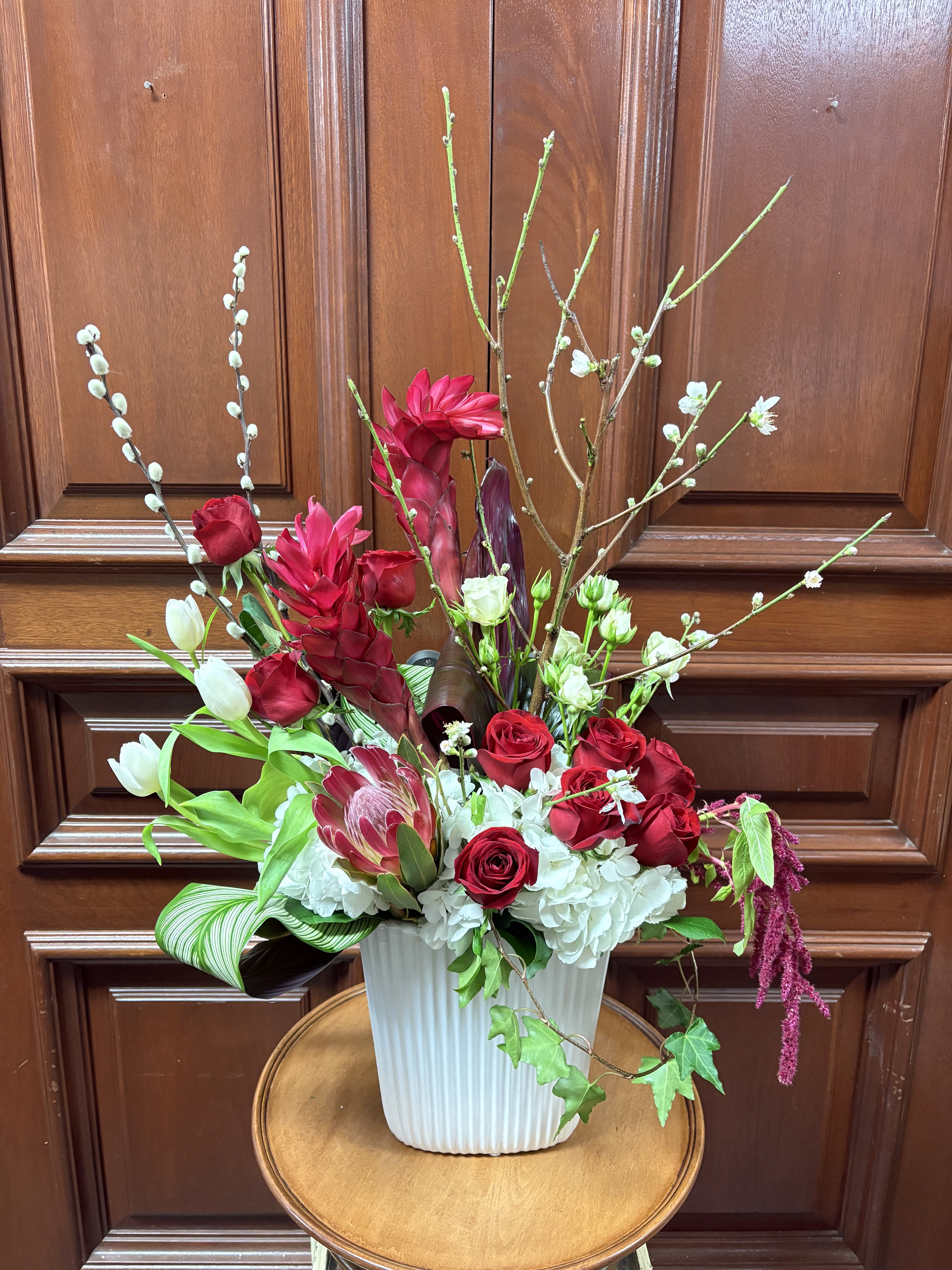 You're so fanciful - Recipe: Seasonal Cherry Blossoms, White Hydrangaes, White Tulips, White Spray Roses, Ginger, Protea, Roses, Red Amarathus, Pussy Willow and Tropical Leaves in a tall, white vase. Seasonal: Seasonal Cherry Blossoms Orientation: Front Facing View Photo shown: Standard