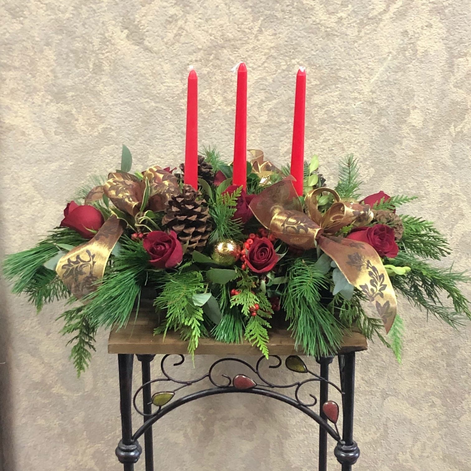 Traditional Pine Centerpiece - Type of Flowers: Red Roses, Pine Cones, Pine greens, and Berries with three red candles, and gold ribbon on a plate. Availability: All year round Substitute Available: Yes Design View: All Facing View Photo shown: Regular 
