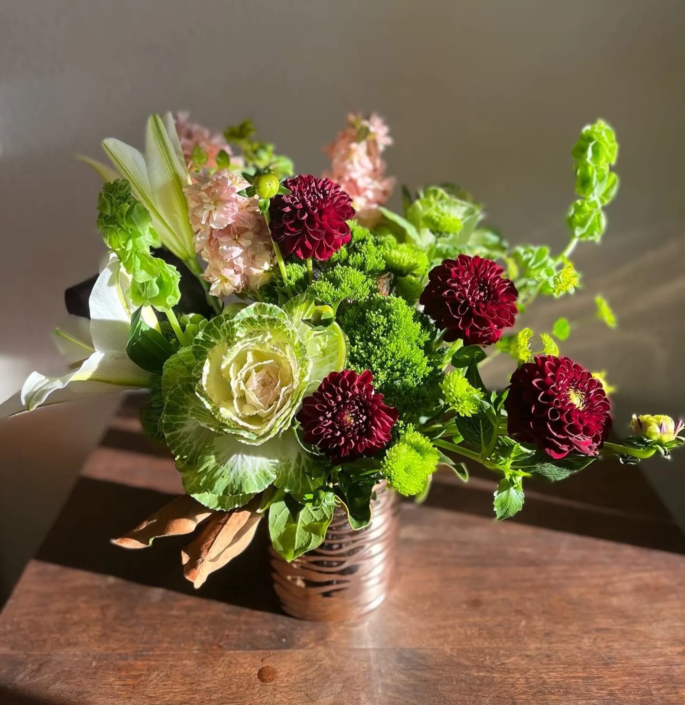 Wine and Rosé  - This designs has shades of greens, reds, and pinks that will bring a glowing smile to someone's face. Type of Flowers: White Lilies, Black Dahlias, Blush Pink Stock, Green Hydrangeas, Bells of Ireland, and Cabbage in a short rose pink vase. Availability: Dahlias available mid-summer to autumn. Design View: Symmetric Front Facing View Photo shown: Regular 