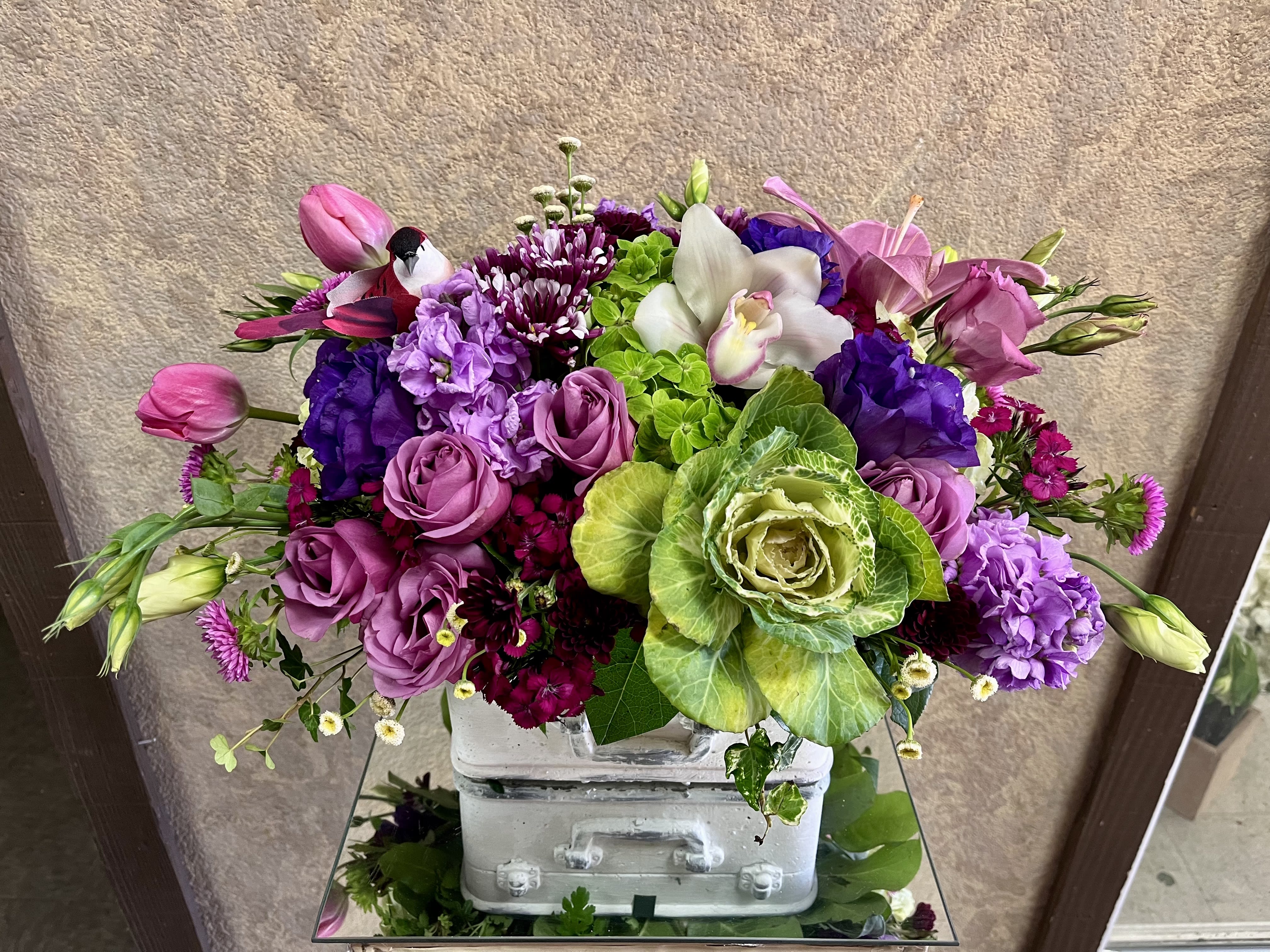 Varenna, Italy - This design is great for birthdays or to say thank you. Recipe: Lavender Stock, Purple Lisianthus, White Cymbidium Orchids, Lavender Roses, Green Hydrangeas, Pink Tulips, Green Cabbage, Chrysanthemums, Matsumoto Asters, Sweet William and more in a white, travel suitcase planter. Availability: All year round Design View: Symmetric Front Facing View Photo shown: Standard