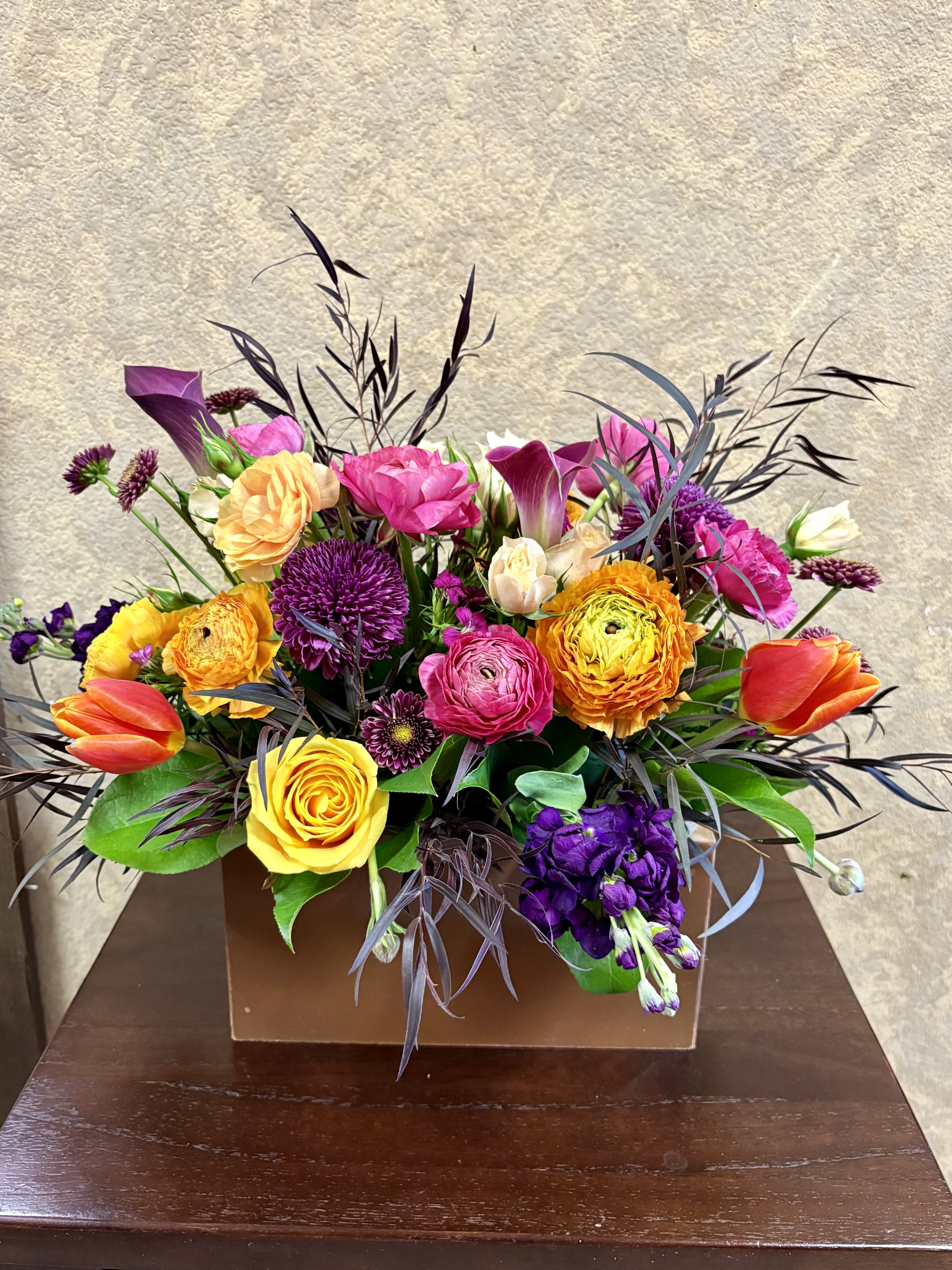 Low rectangular arrangement of bright yellow, orange, pink, and purple flowers in a brown box.