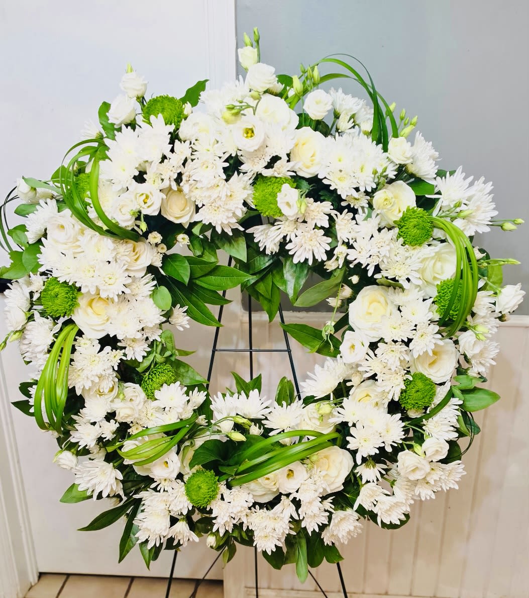 Serenity Everlasting  - Pure white blossoms arranged in a perfectly round tribute-this Sympathy and Funeral wreath offers a calm, ceremonial presence that helps the person you're honoring feel remembered with grace. The Serenity Everlasting flower arrangement reads as a formal, peaceful salute: the recipient will sense care and respect from its clean white-and-green palette and balanced, symmetrical design.  Dense clusters of chrysanthemums, soft lisianthus, and white roses form the full face of the wreath, accented by bright green button centers and looping green foliage for contrast and texture. Mounted on a black wreath stand, the arrangement is substantial and polished-ideal for memorial services, graveside displays, or a church setting. The circular shape symbolizes continuity and remembrance while the serene colors convey compassion and quiet dignity.  Choose this wreath when you want to express sympathy, honor a life well lived, or mark a funeral with a calm, respectful tribute. Arrives ready on an easel-style wreath stand suitable for display; for local delivery and pickup in West Frankfort, IL, we'll handle careful placement so the person you're thinking of is seen with the dignity they deserve.
