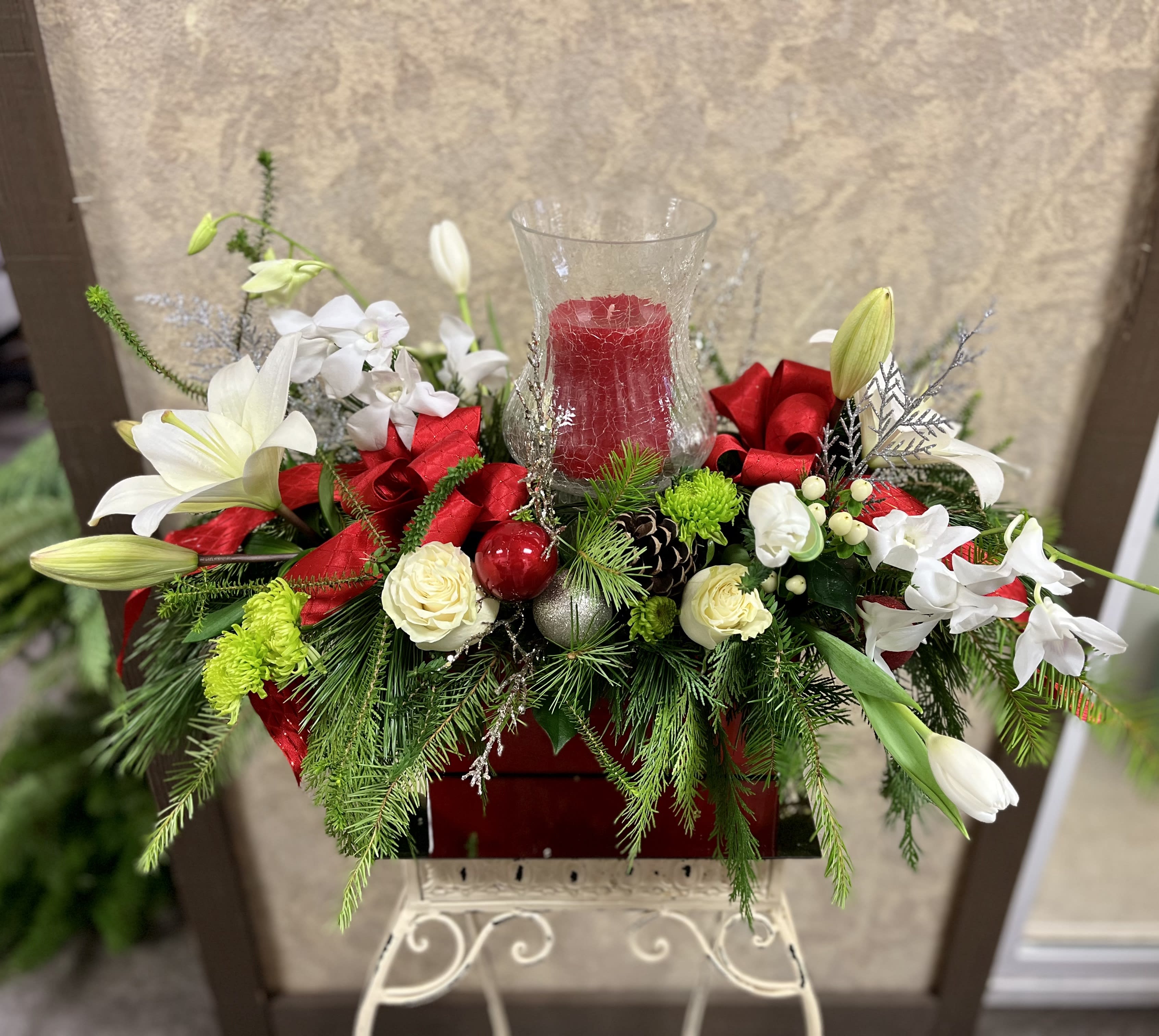 Holly Jolly Christmas Centerpiece - Type of Flowers: White Lilies, Orchids, Roses, Tulips, Green Buttons with a candle, ornaments, ribbon, pinecones, and more in a red rectangular vase. Design View: All Facing View Photo shown: Regular 