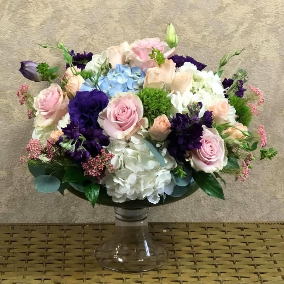 You Swept Me Away Centerpiece - Type of Flowers: White and Blue Hydrangeas, Purple Stock, Purple Lisianthus, Pink Roses, Blush Spray Roses, Dianthus Barbatus, and Pink filler in a urn crystal vase. Availability: All year round Substitute Available: Yes Design View: All Facing View Photo shown: Regular