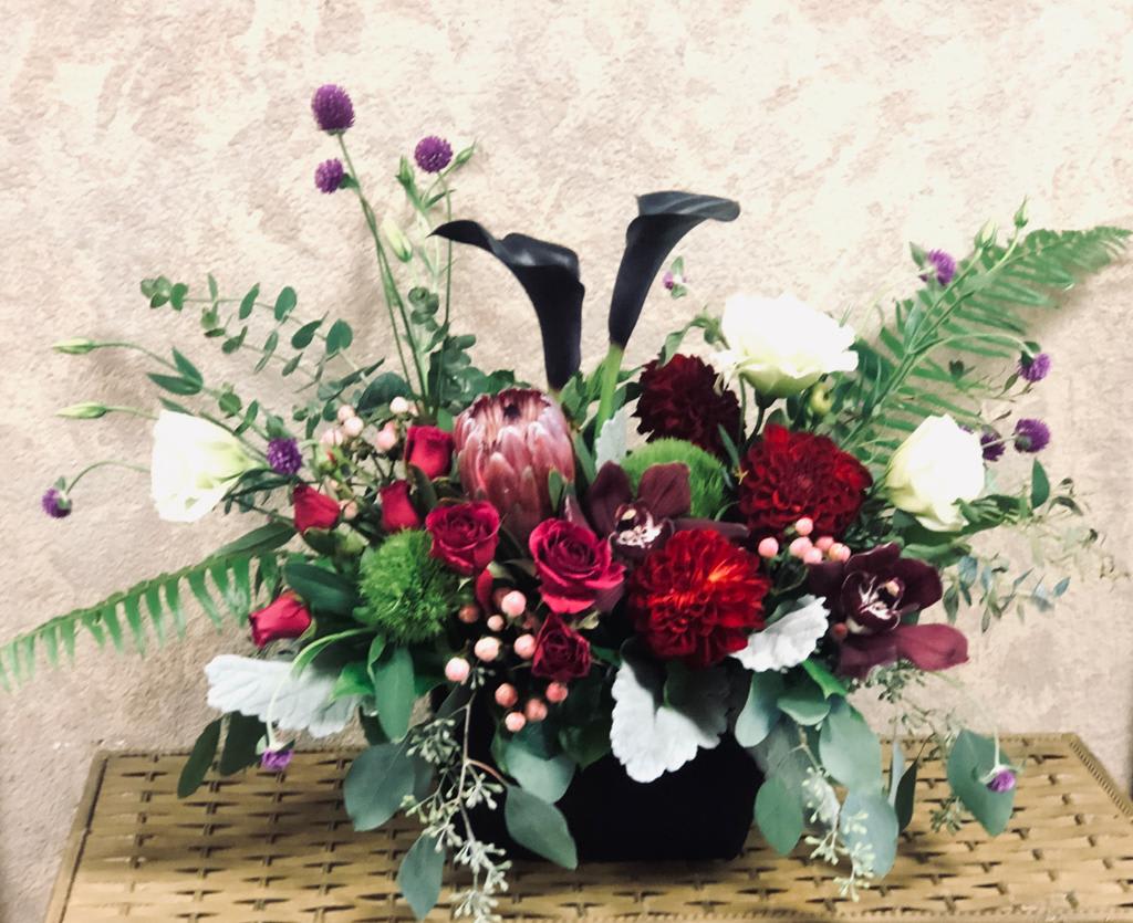 Tango en la Calle 13 - Type of Flowers: White Lisianthus, Red Dahlias, Pink Protea, Red Spray Roses, Black Calla Lilies, Purple Cymbidium Orchids, Globe Amaranths, Dianthus Barbatus, Hypericum Berries, Dusty Miller and Greens in a black, ceramic vase. Availability: All year round Substitute Available: Yes Design View: All Facing View Photo shown: Regular 