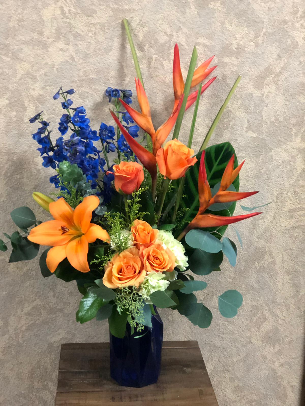 Formentera, Ibiza, Spain - Type of Flowers: Orange Birds of Paradise, Blue Delphinium, Orange Roses, Orange Asiatic Lilies, White Hydrangea, and greens in a blue vase. Availability: All year round Substitute Available: Yes Design View: All Facing View Photo shown: Regular 